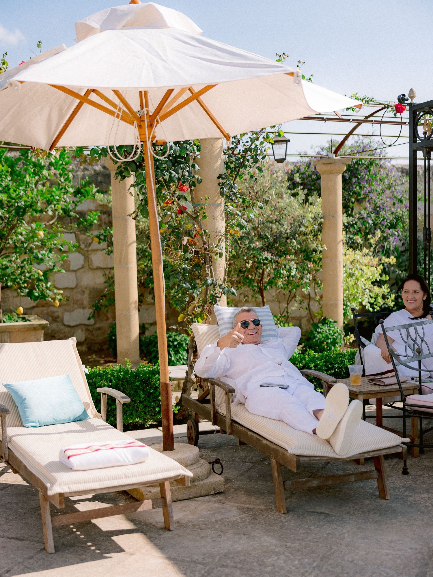 Outdoor lounge area with wooden chaise lounges, cream umbrellas and stone columns in a peaceful garden setting.