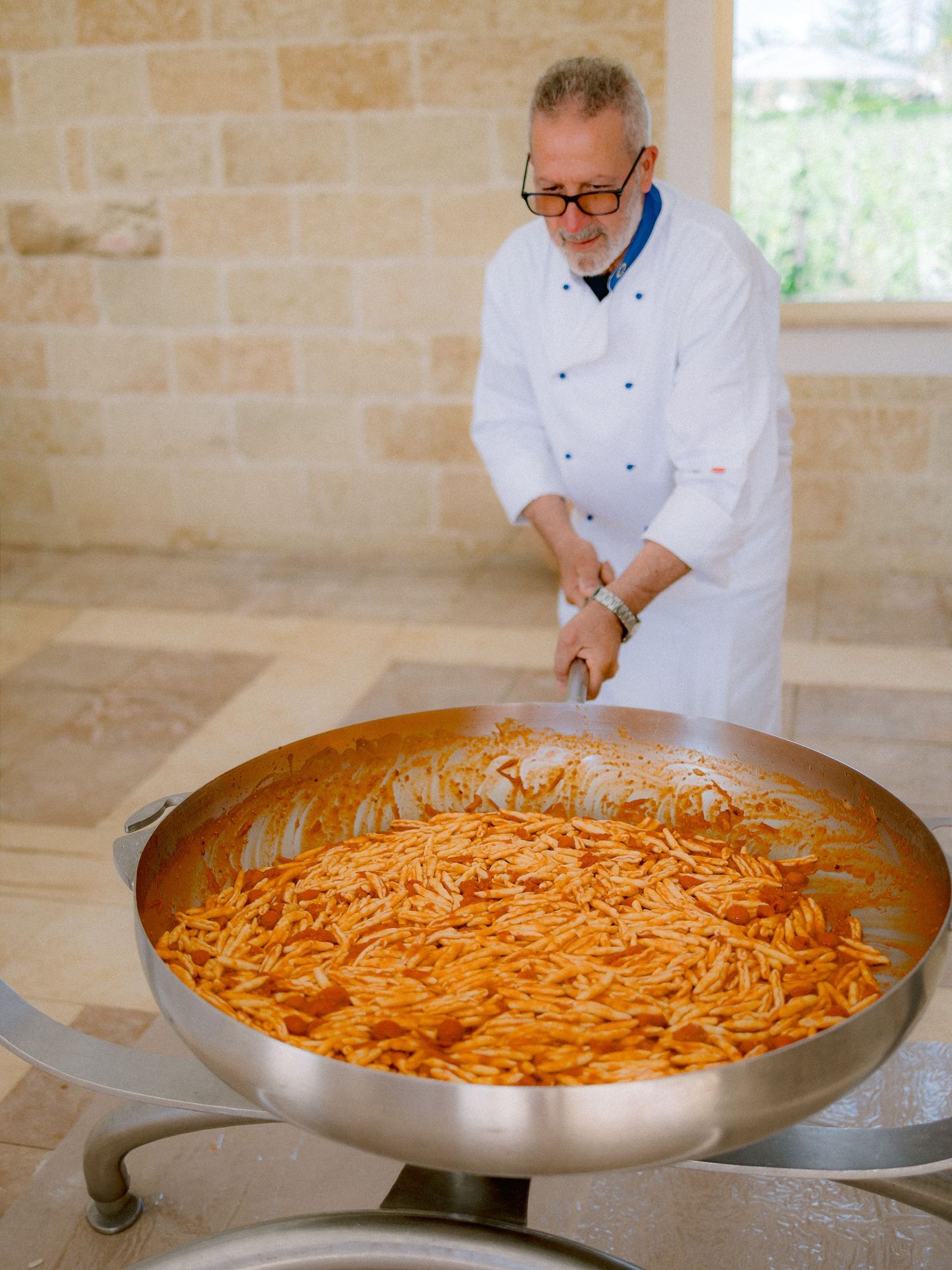 A chef in white uniform prepares a large pan of pasta with tomato sauce in a professional kitchen with stone walls.