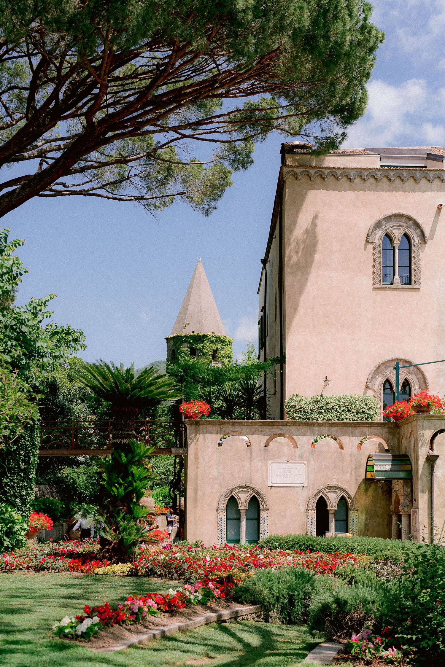 Villa Cimbrone in Ravello with pink facade, arched windows, and tower surrounded by lush gardens with red flowers and pine trees.