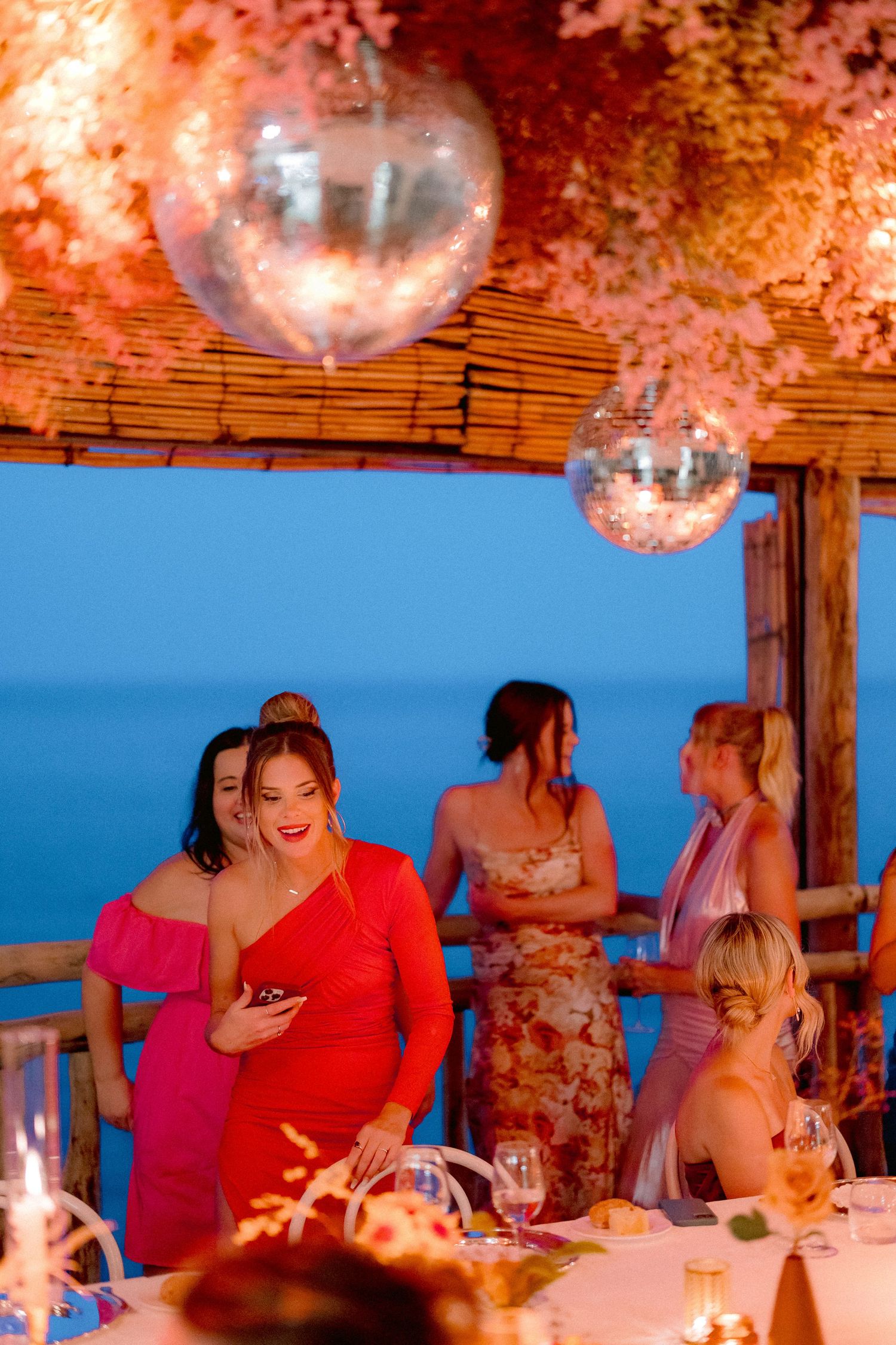 Pink and warm-toned evening scene at an oceanfront restaurant with disco balls hanging from wooden beams and dramatic lighting.