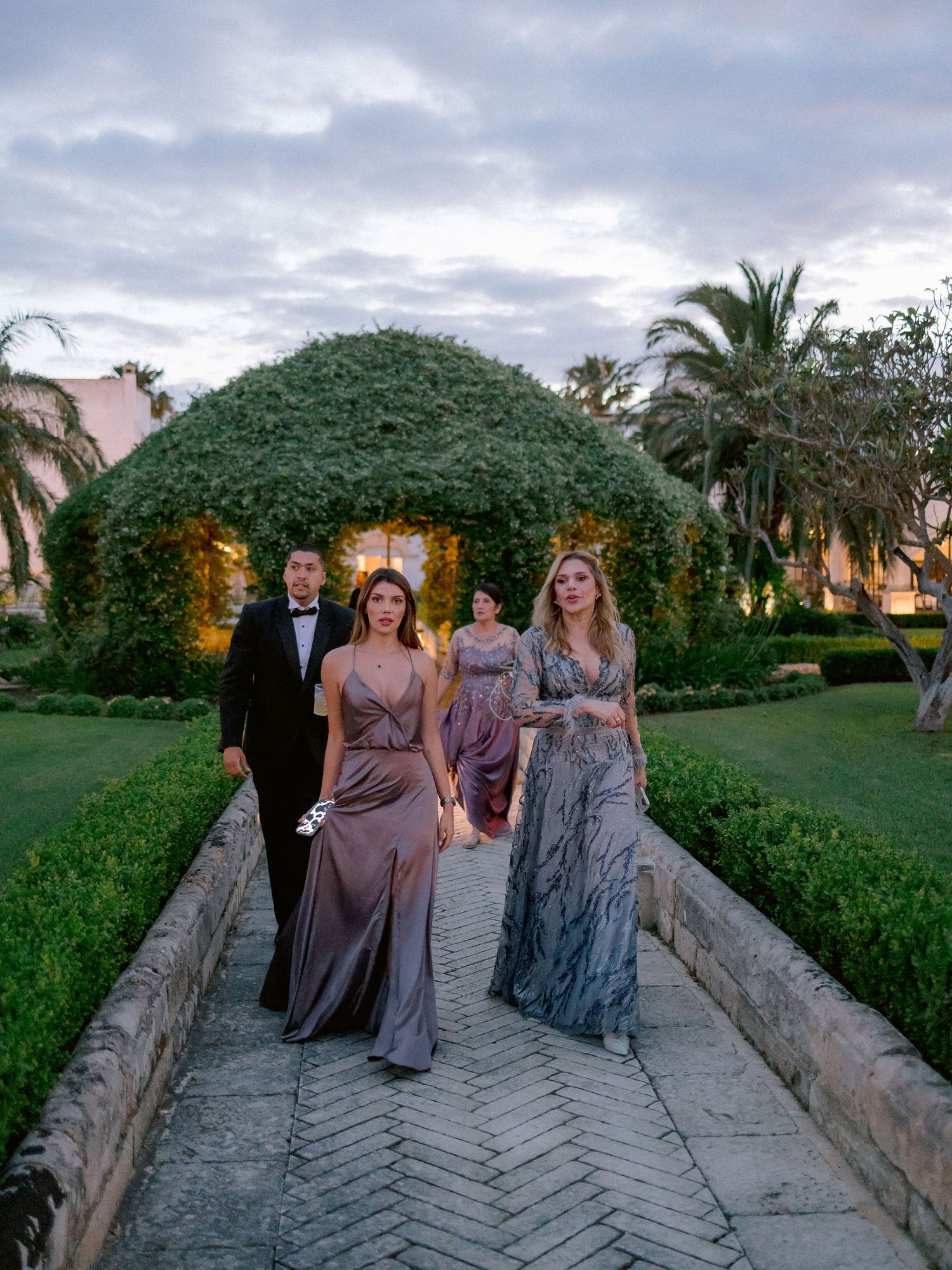 Group in elegant formal attire walking along a garden path with illuminated topiary arch and palm trees at dusk.