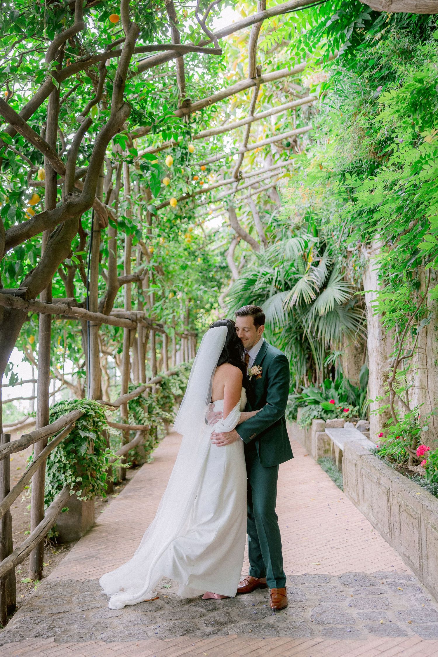 A couple in wedding attire embraces under a lush garden pergola with greenery and climbing vines overhead.