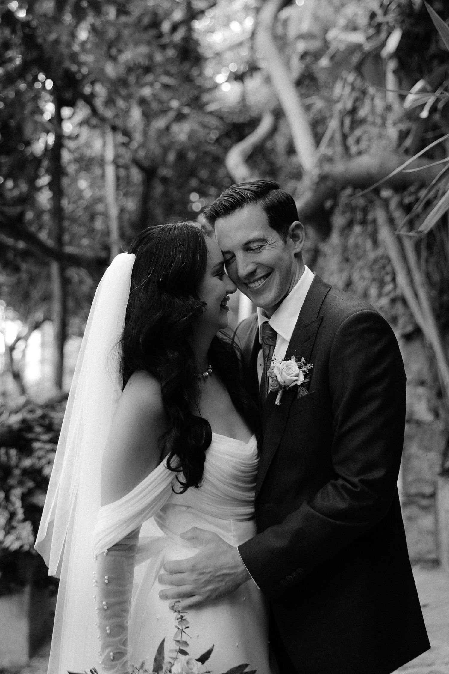 A black and white wedding photograph showing a tender moment between a bride and groom in an outdoor garden setting.