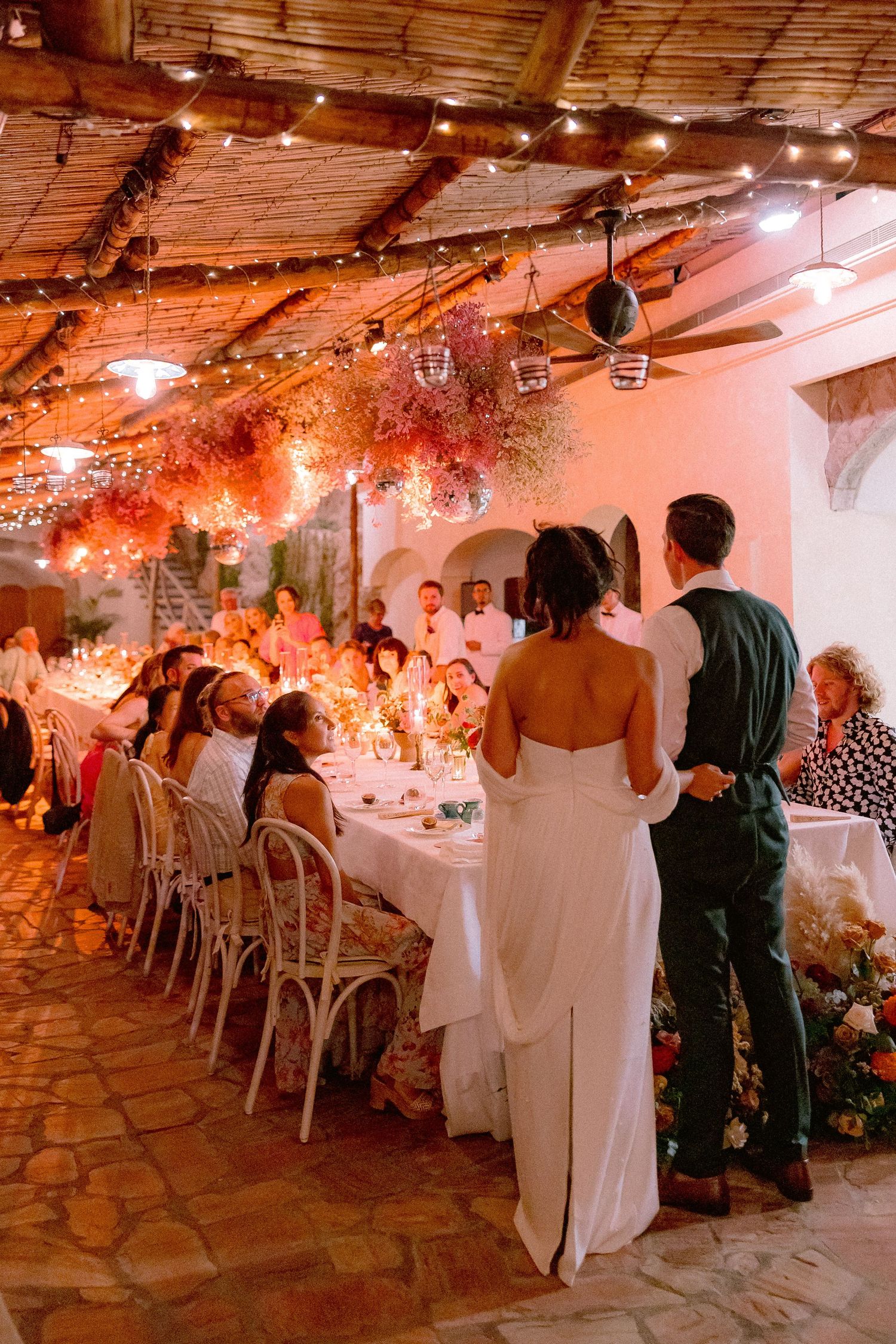 Wedding reception in rustic venue with string lights and floral decor as guests gather around a long table for dinner.