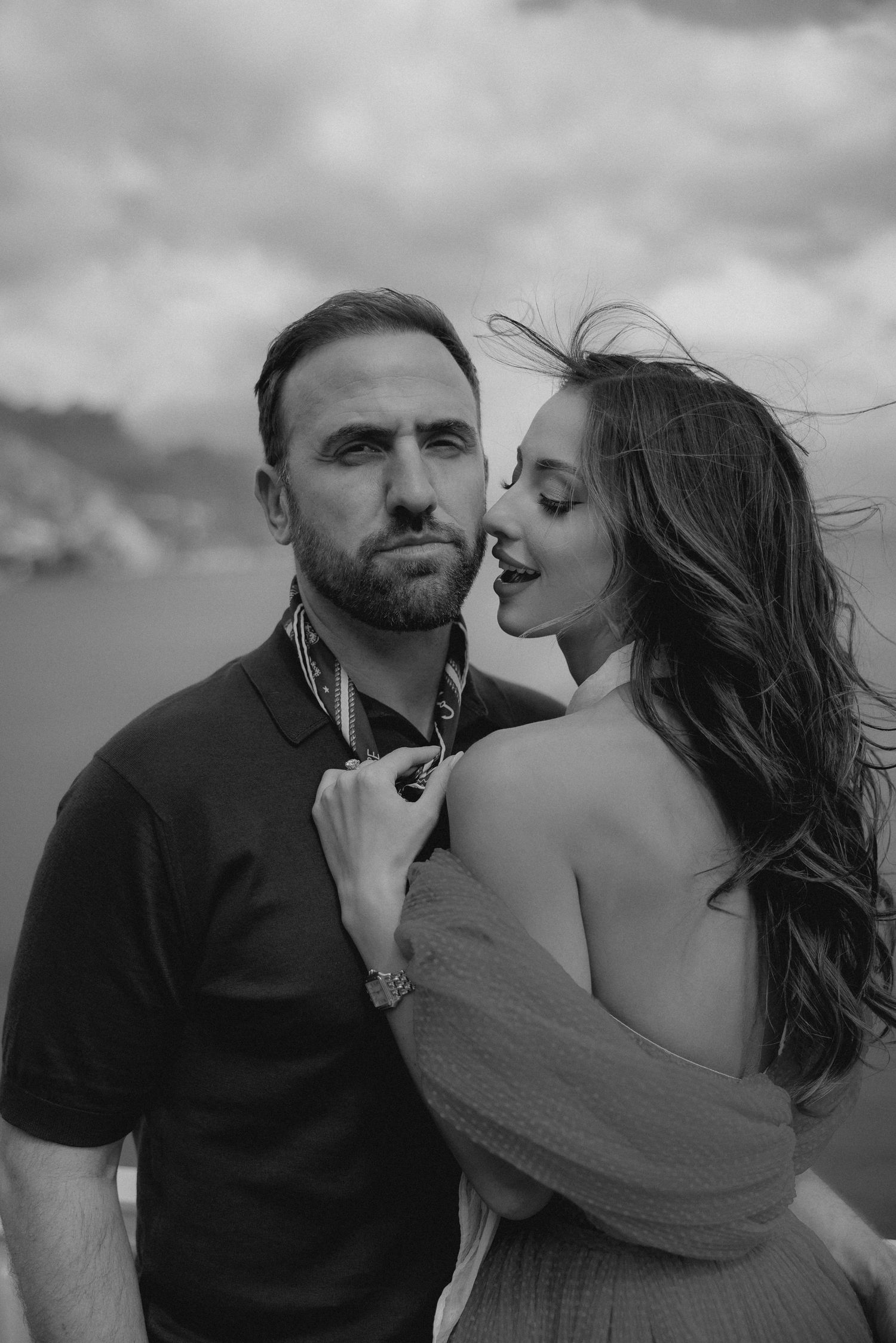 A romantic black and white portrait of a couple embracing outdoors with wind-blown hair and dramatic lighting.