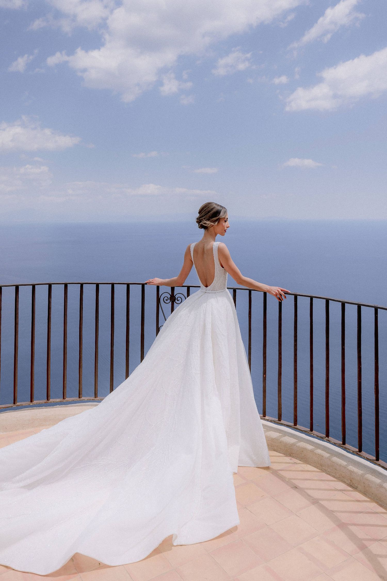 A bride in an elegant white backless wedding gown stands at a scenic coastal overlook with a flowing train at Terrazza dell'Infinito.