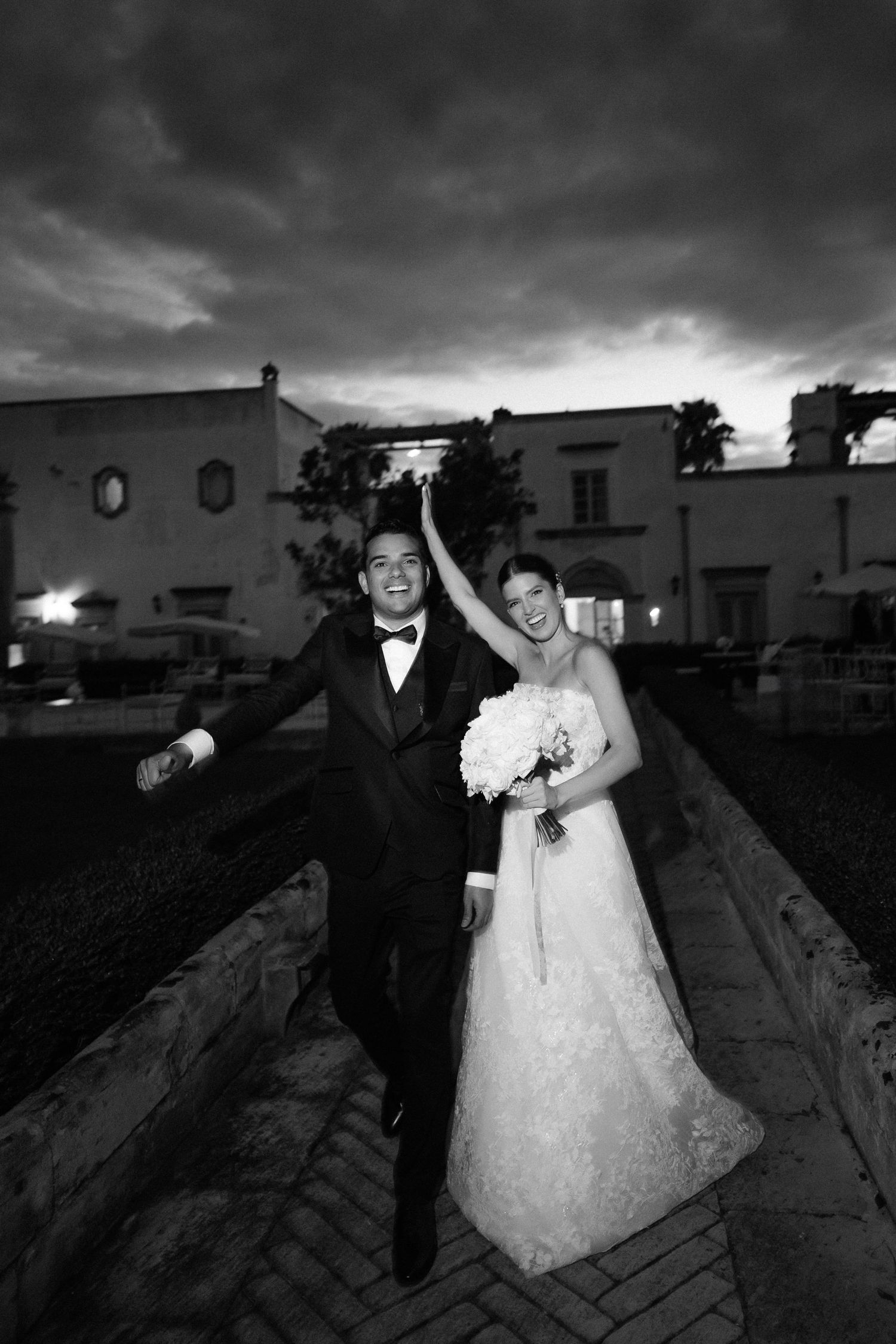 A joyful black and white wedding photo showing a couple celebrating outdoors at dusk with dramatic storm clouds overhead.