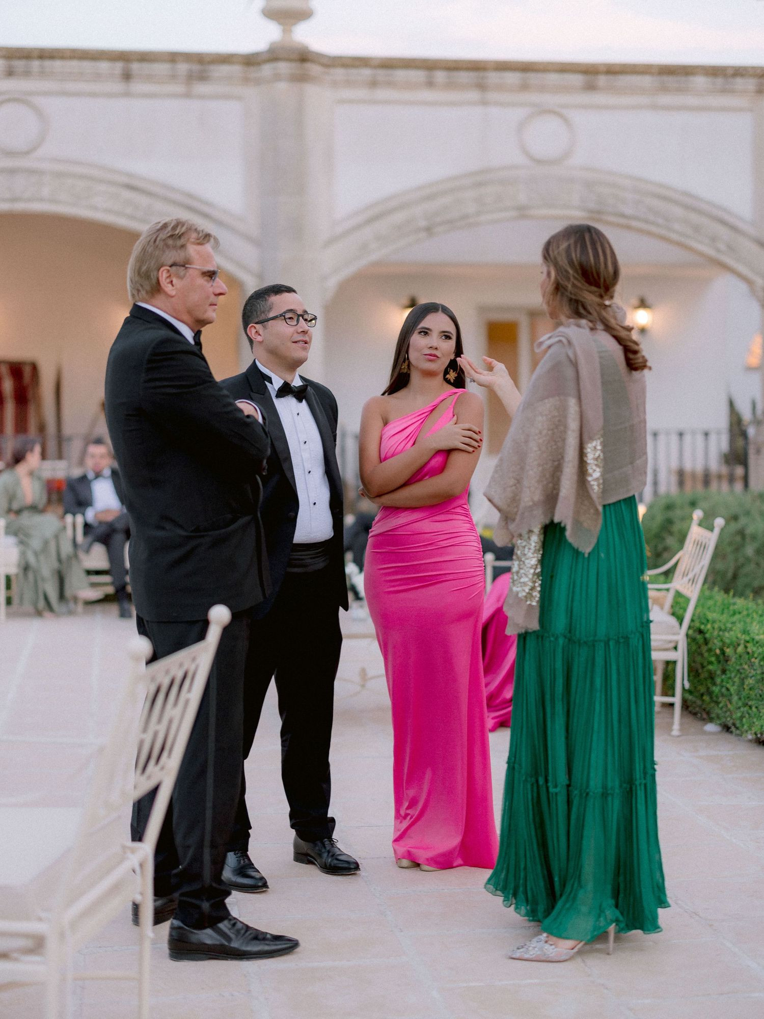 A group of formally dressed people conversing at an elegant outdoor venue with arched architecture in the background.