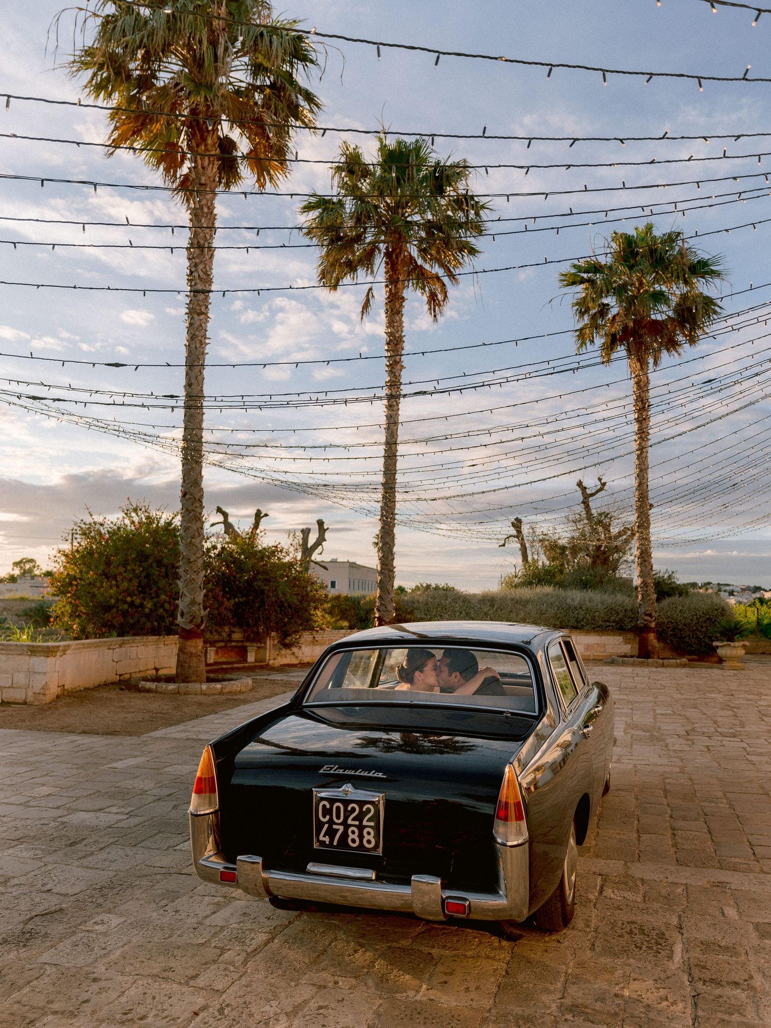 Vintage black car parked beneath palm trees and string lights at sunset with Mediterranean coastal scenery in background.