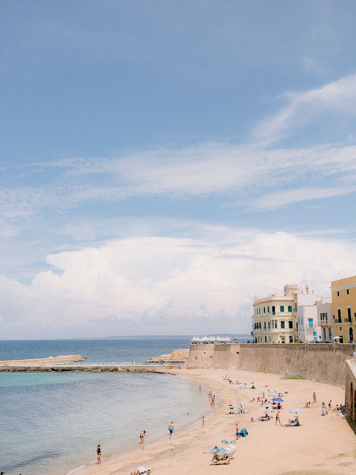 Scenic Mediterranean coastal beach in Gallipoli with beachgoers relaxing beneath blue skies and historic buildings along the shoreline.