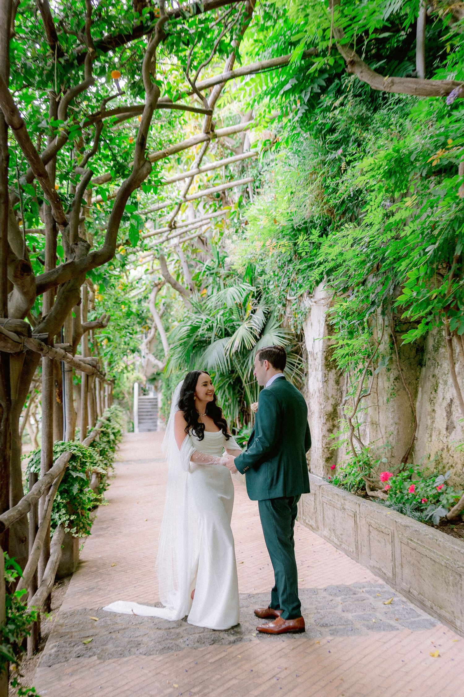 A couple shares a romantic moment on a garden path lined with lush green vines and trees.