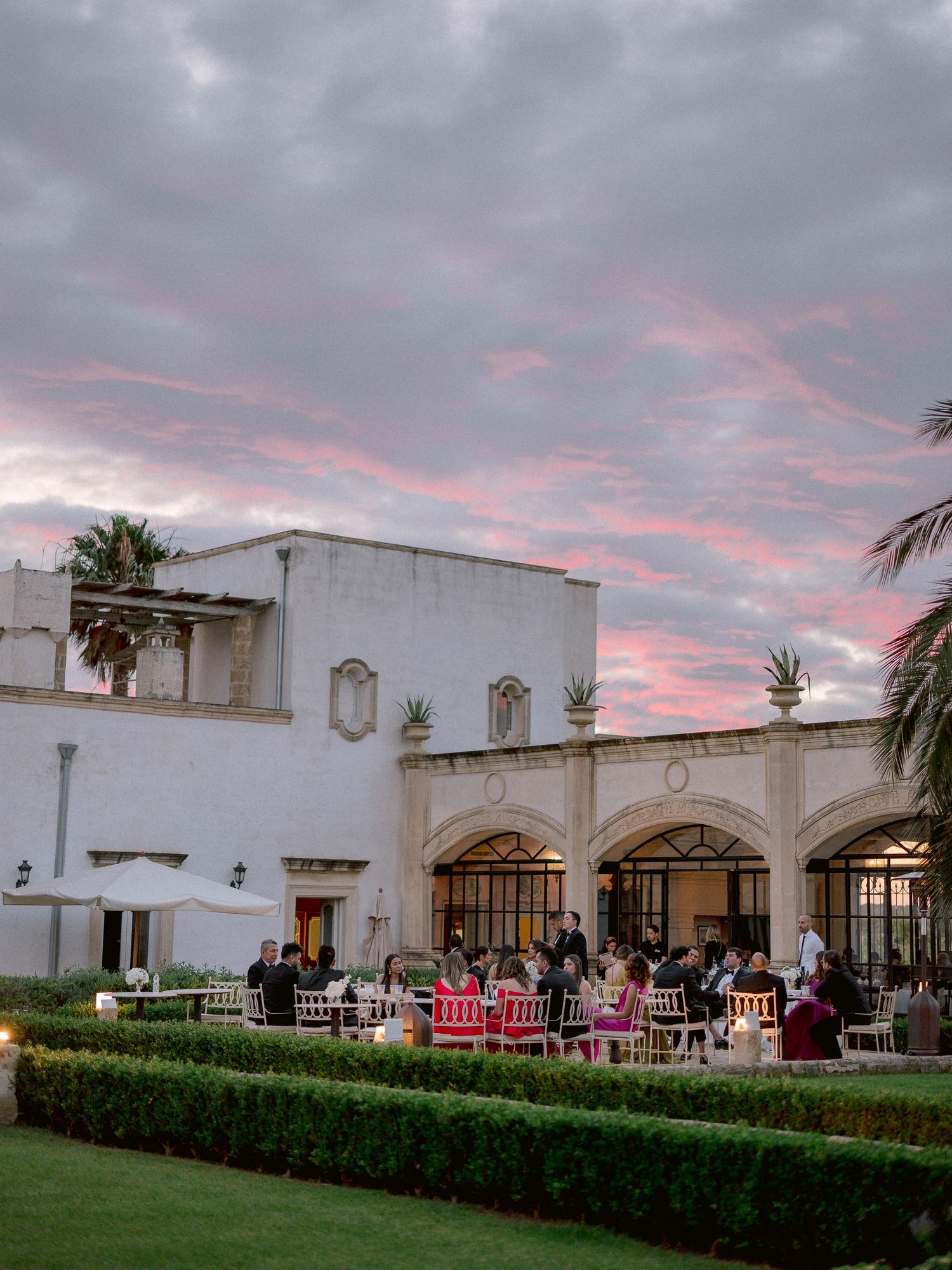 White Spanish-style venue with arched windows and outdoor seating area under pink sunset clouds during an evening event.