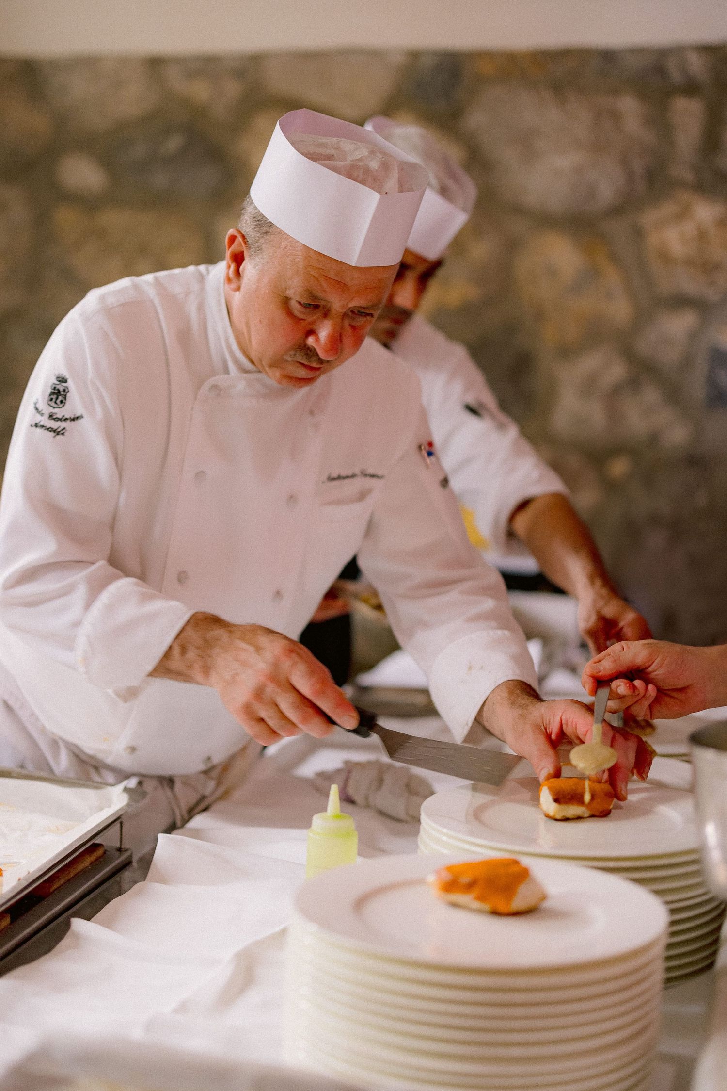 A chef in traditional white uniform and hat carefully plates dishes in a professional kitchen setting next to stacked plates.