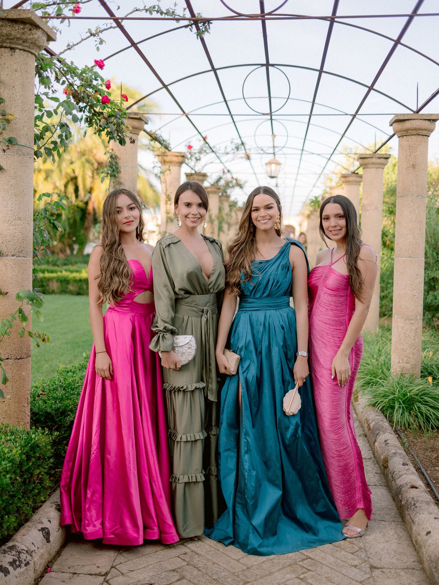 Four women wearing elegant evening gowns in pink, olive, teal and rose stand under a covered garden walkway with columns.