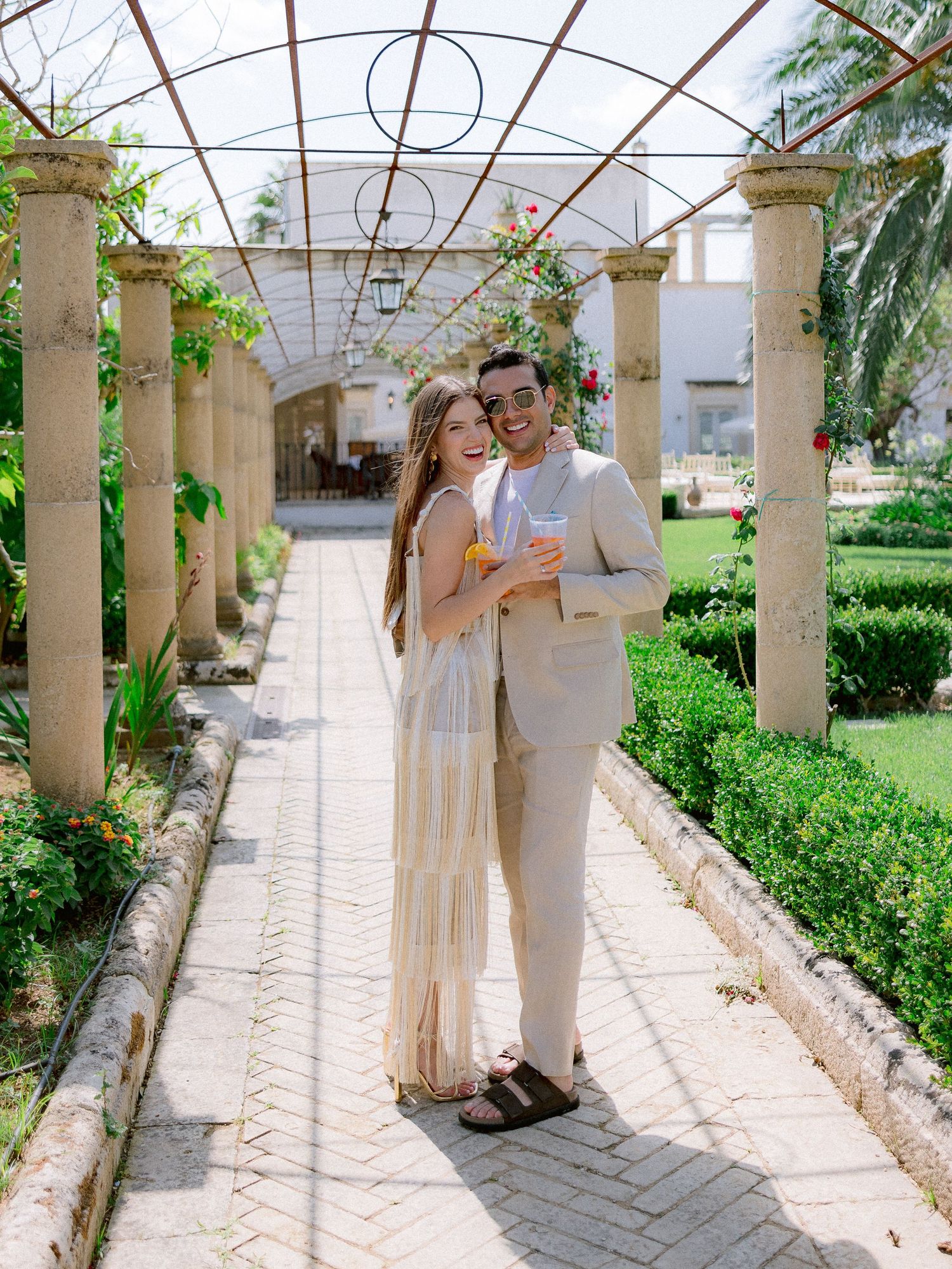 A couple stands affectionately in a garden pergola walkway with stone columns and tropical greenery.