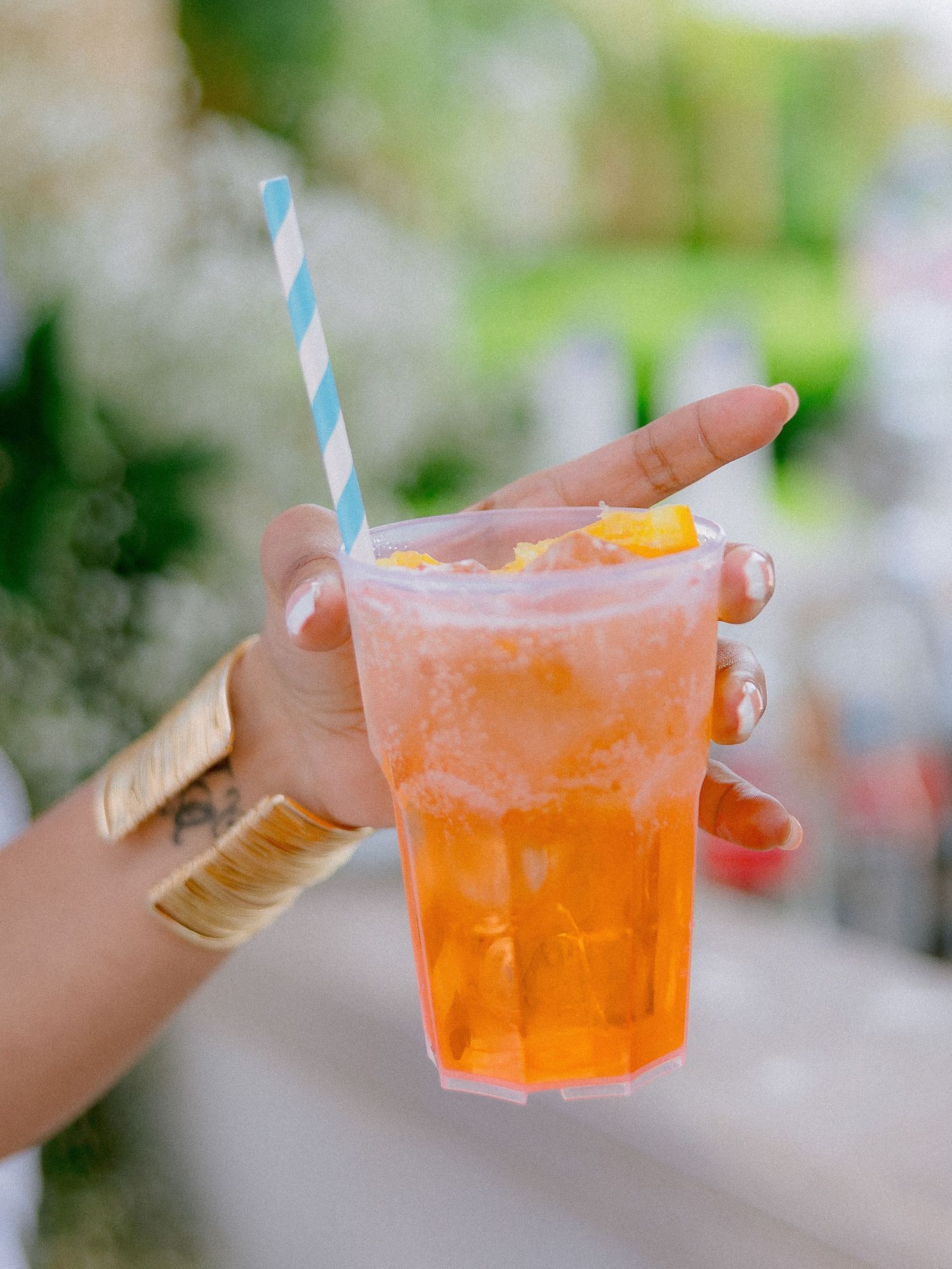 Hand holding a refreshing orange cocktail with ice and blue striped straw in a plastic cup outdoors.