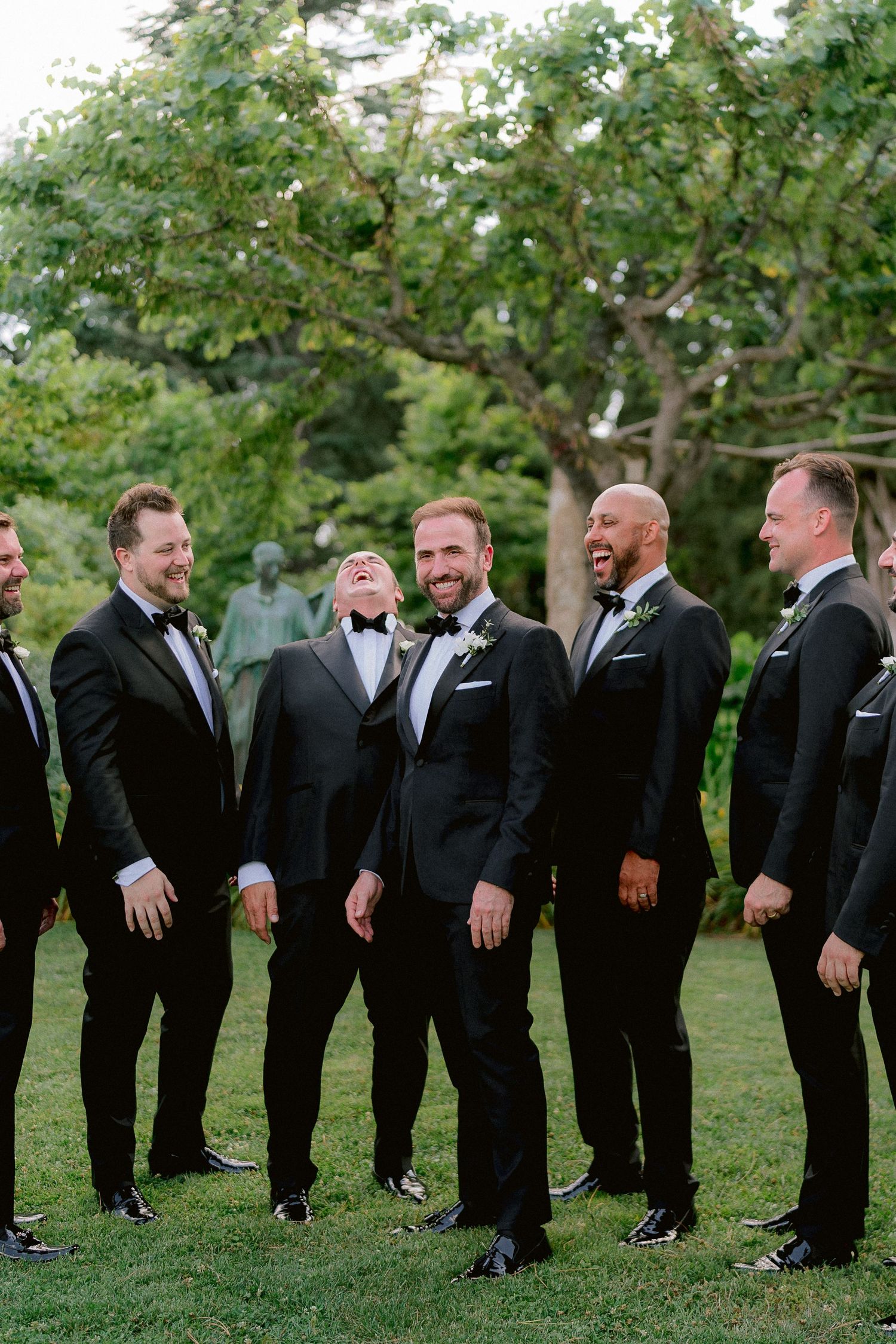 A group of groomsmen in black tuxedos share a candid moment and laugh together on a grassy lawn.