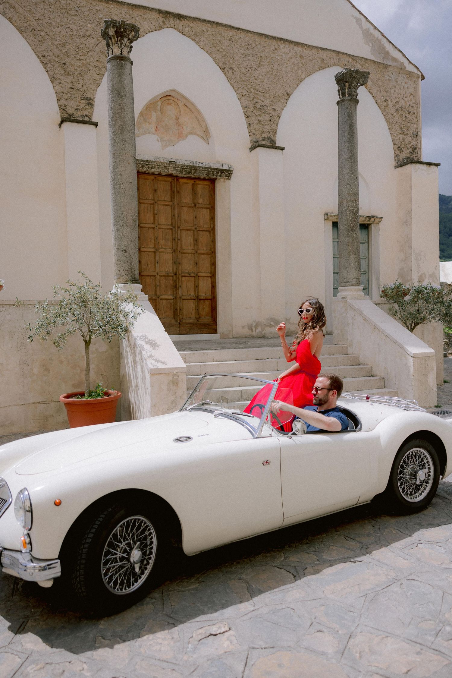 White vintage MG roadster parked in front of Mediterranean church entrance with stone columns and arches.