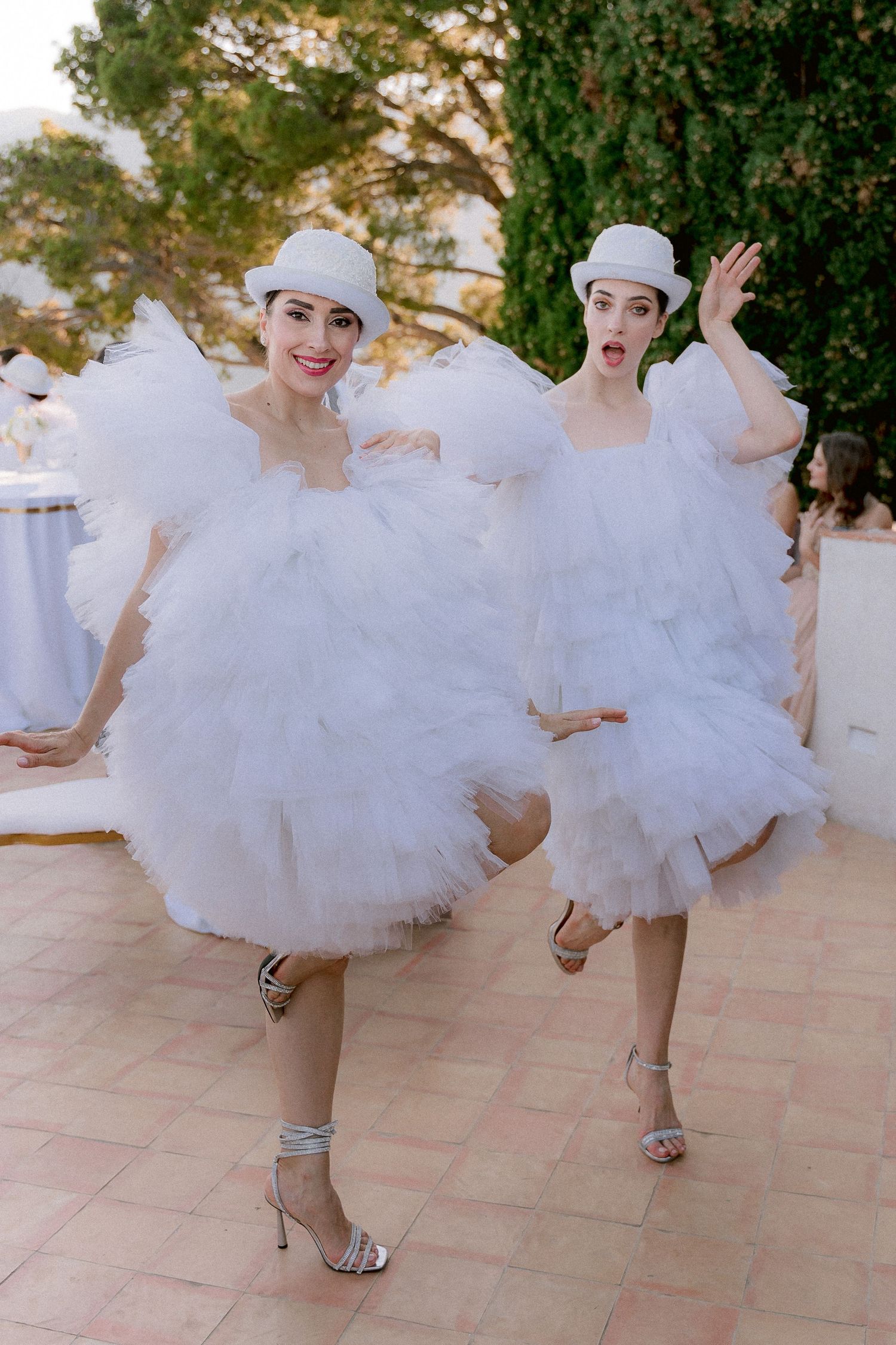 Two dancers in fluffy white tulle dresses and matching caps strike playful poses on an outdoor terrace.