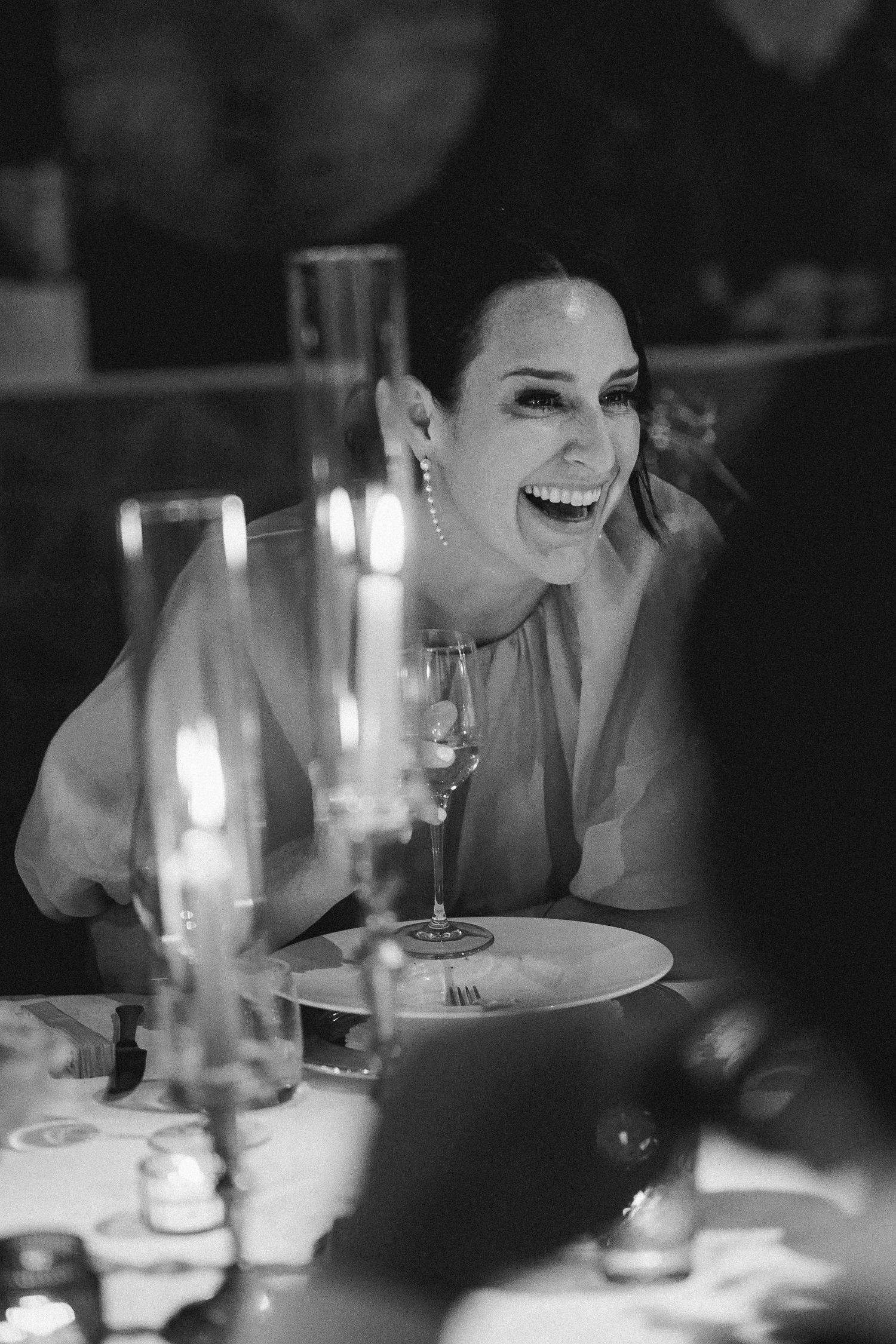 Black and white candid photograph of laughter and joy at an intimate dinner party with wine glasses and candlelight.