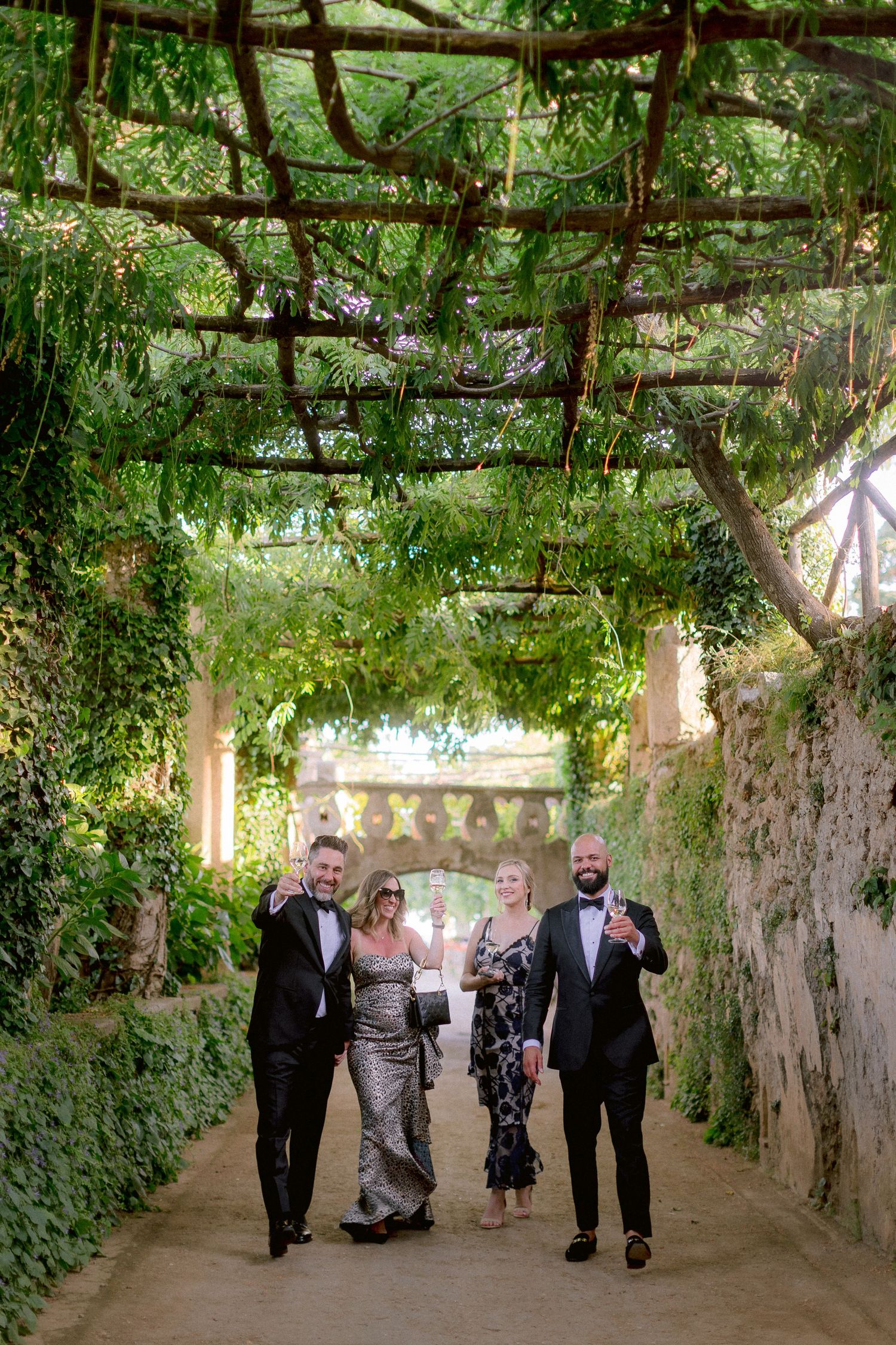 A group walks together along a charming garden path covered by a natural green vine archway in a Mediterranean setting at Villa Cimbrone.
