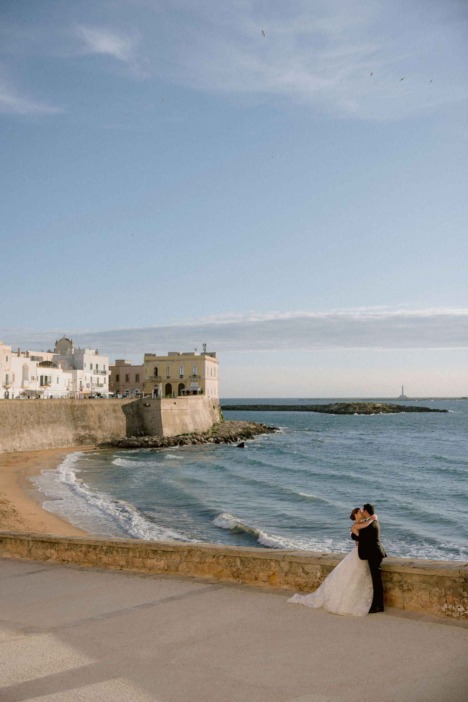 A couple embraces on a stone wall overlooking a scenic Mediterranean beach with historic fortress buildings in Gallipoli, Puglia.