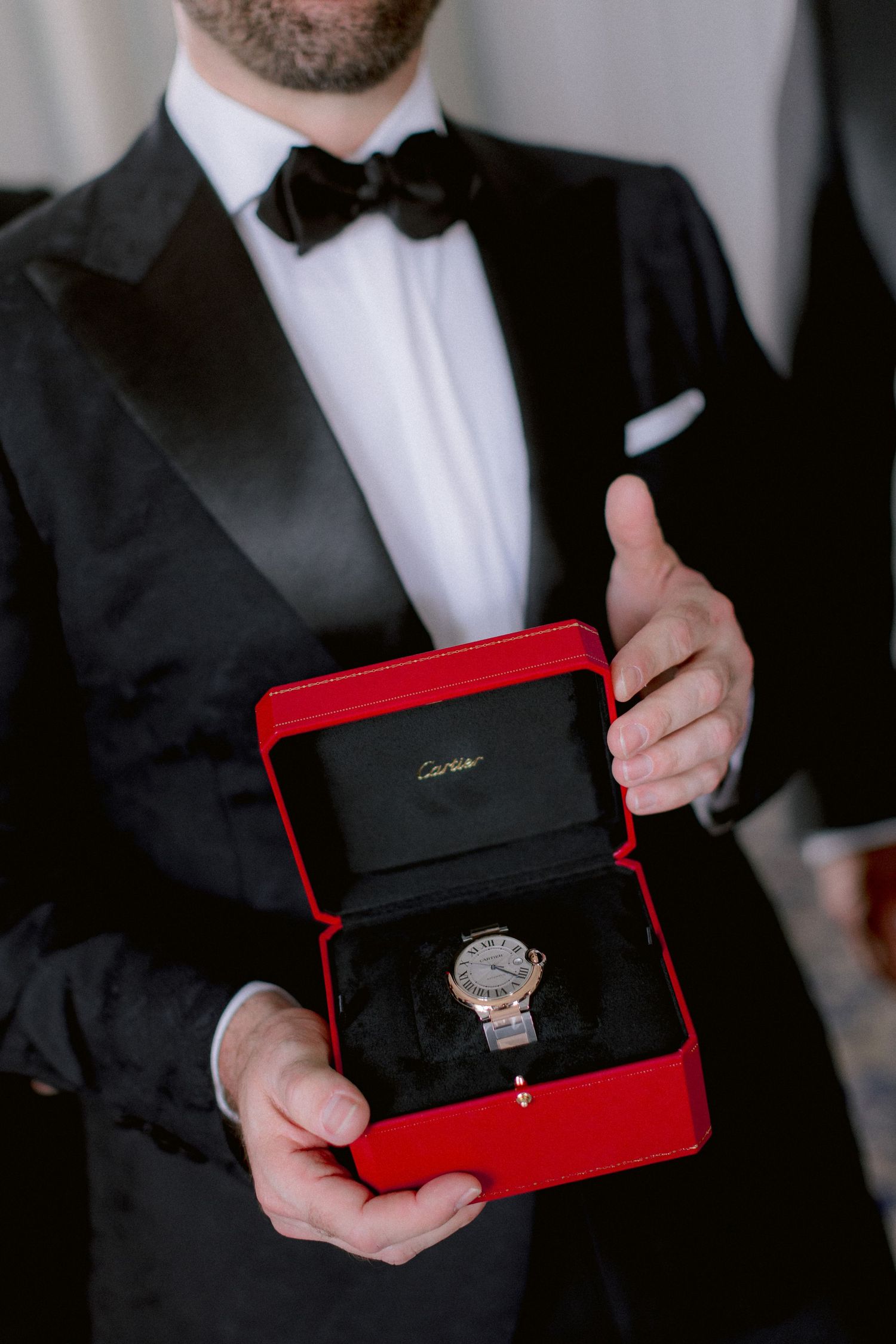 Elegant watch in red Cartier box held by someone wearing formal black tuxedo and bowtie.