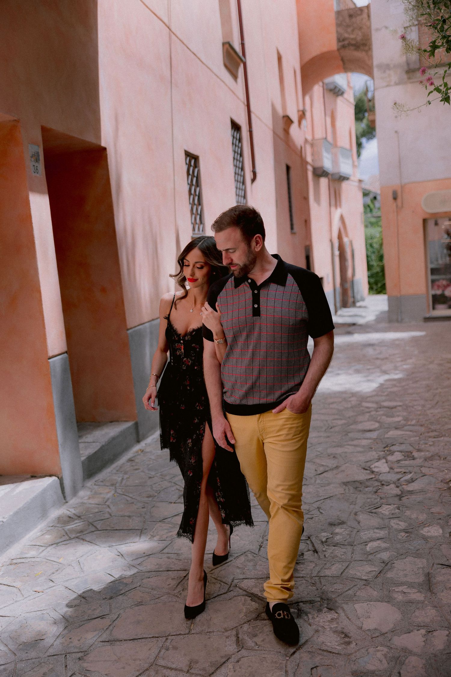 A couple walks together down a quaint street with pink walls and stone pavement in a romantic European setting.