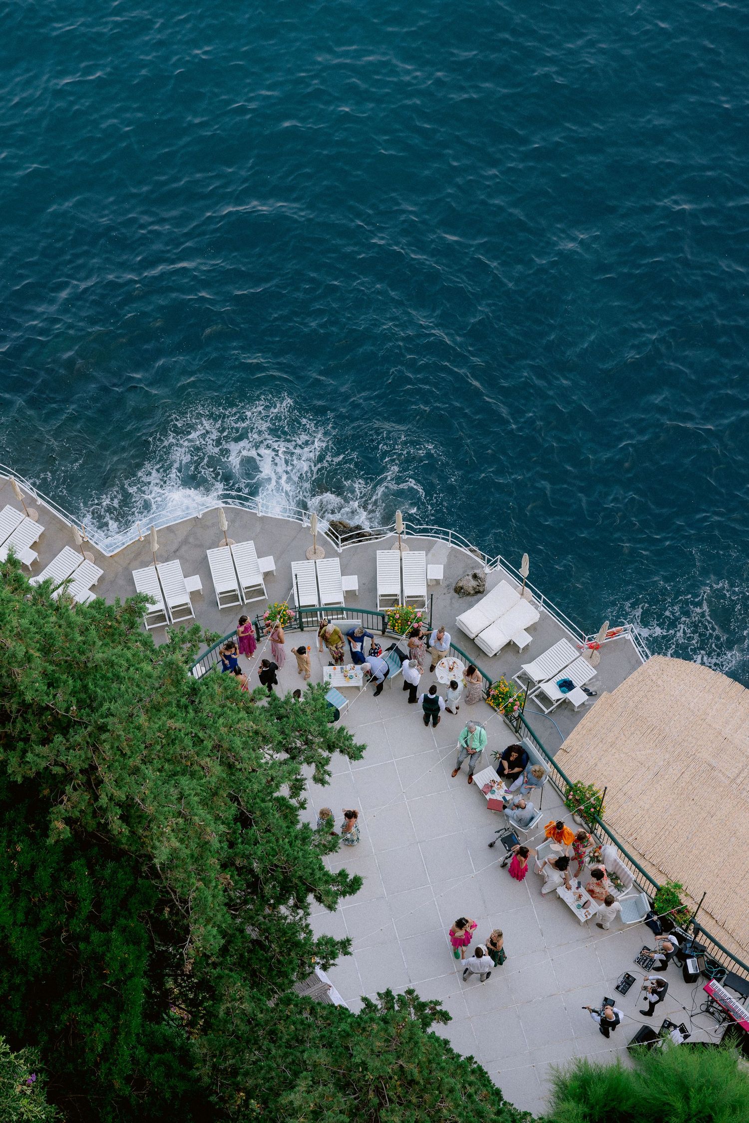 Aerial view of a seaside terrace with outdoor dining tables overlooking deep blue Mediterranean waters in Italy.