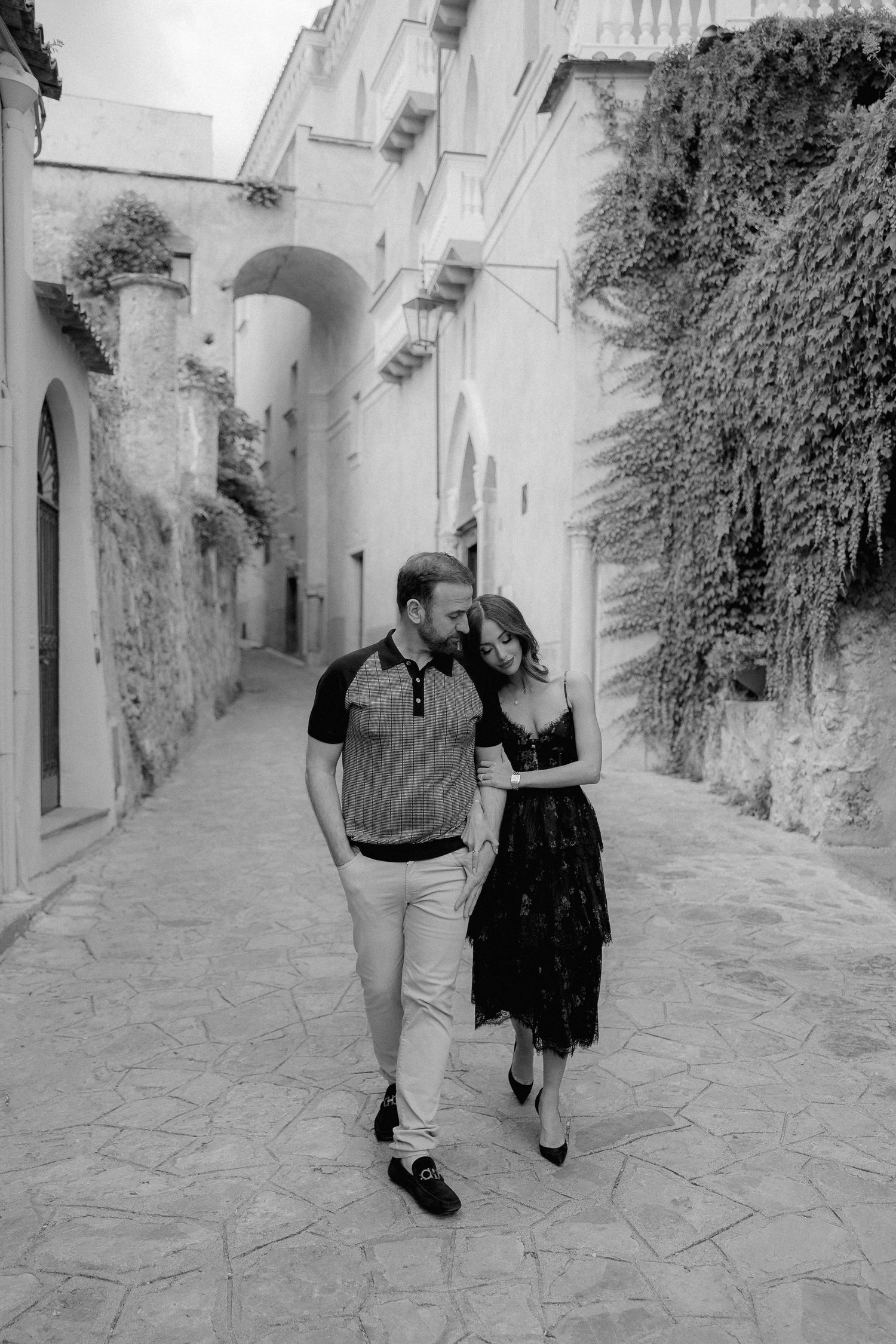 A couple walks together down a charming stone alleyway in an old European town with ivy-covered walls.