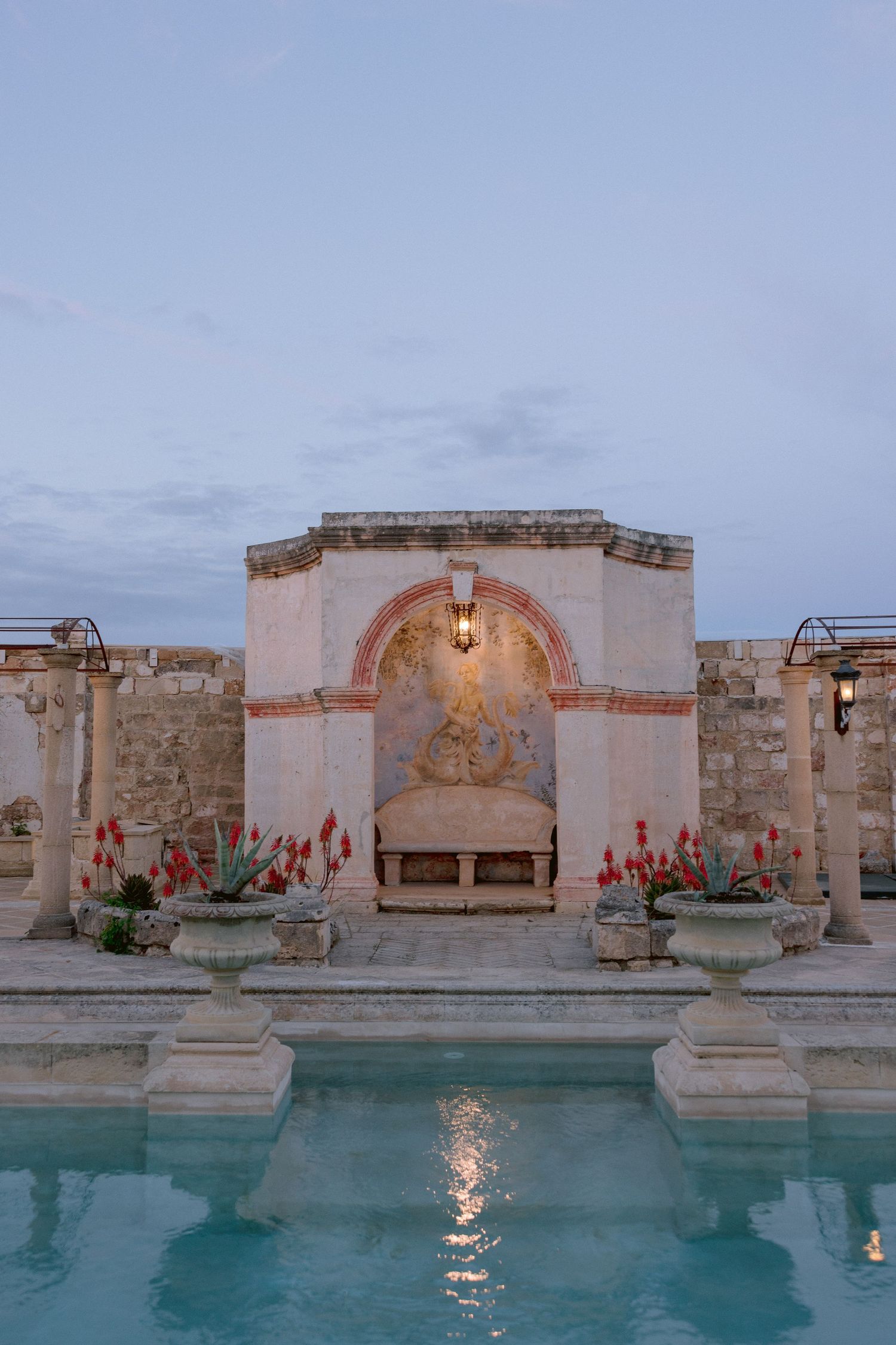 Elegant stone archway with glowing lamp and ornate urns reflected in turquoise pool at dusk.