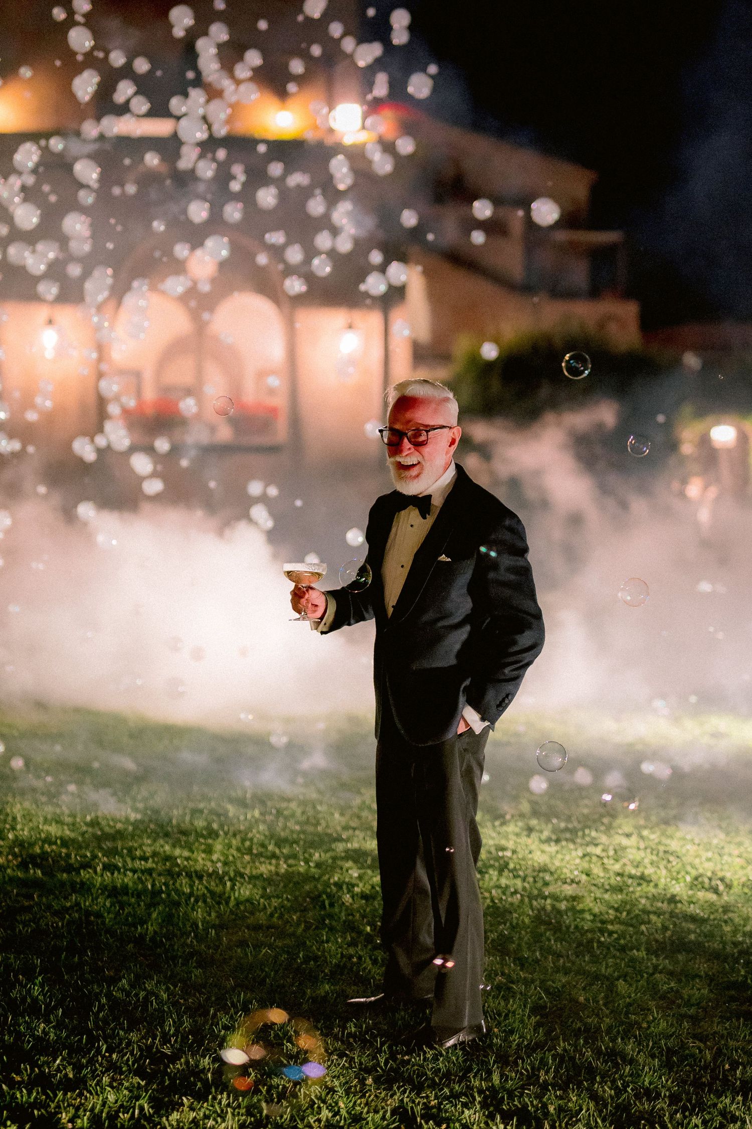 A person in a black tuxedo stands on a lawn at night surrounded by bokeh lights and fog effects.