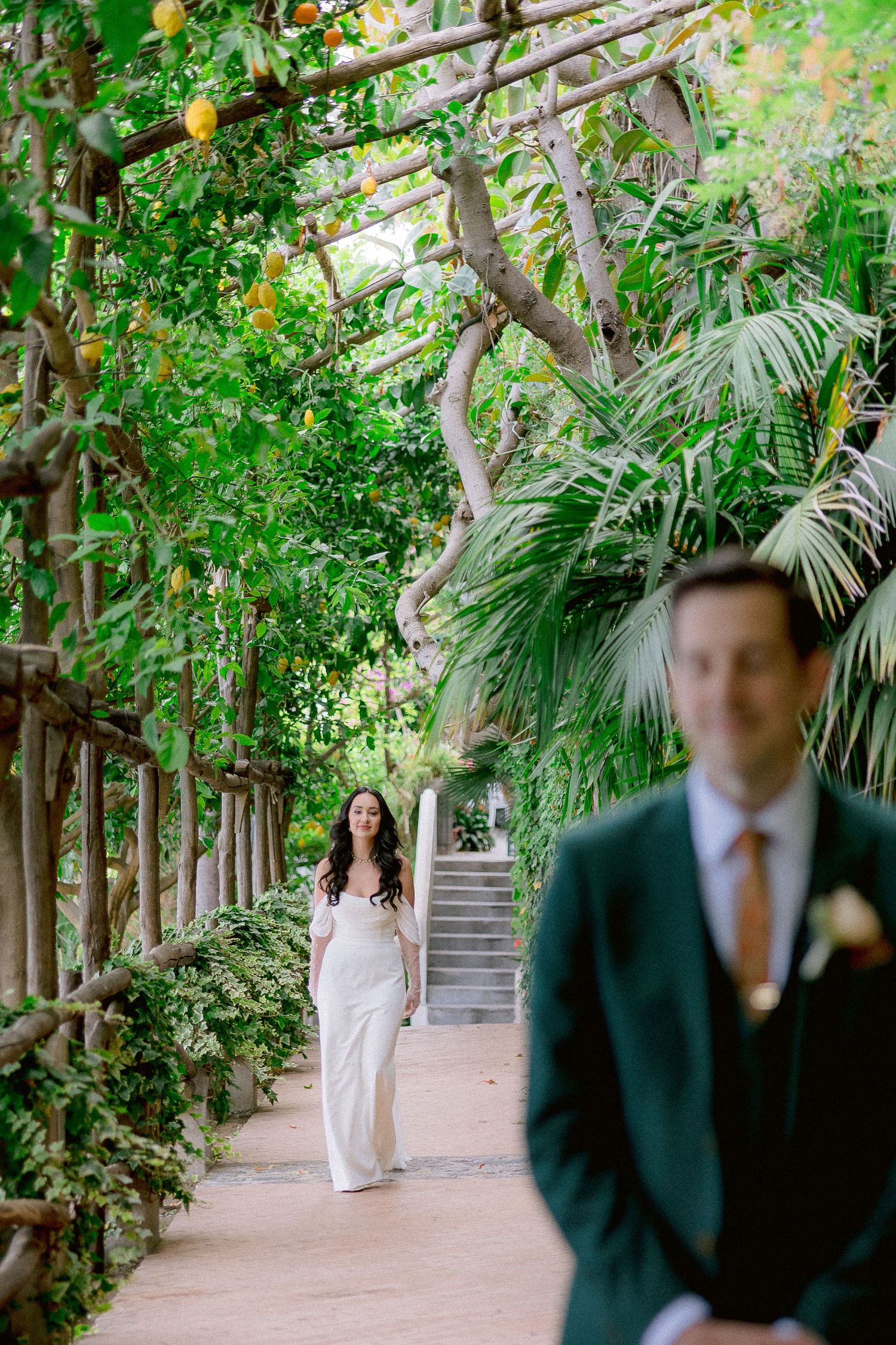 A couple photographed in a lush garden pathway with climbing vines and palm fronds creating a romantic tropical setting.