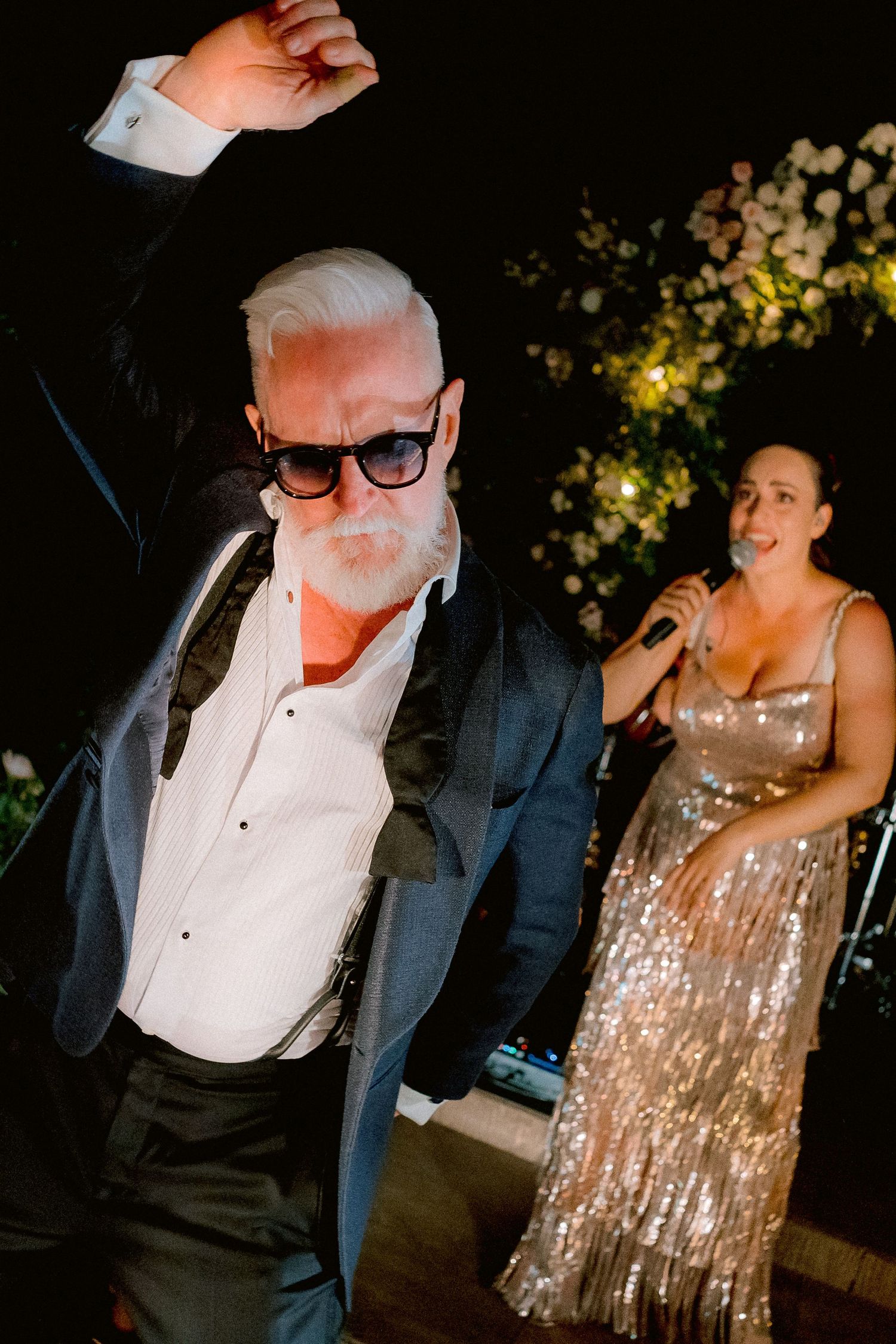 Well-dressed party goers dancing at a nighttime celebration with festive lights and decor in the background.