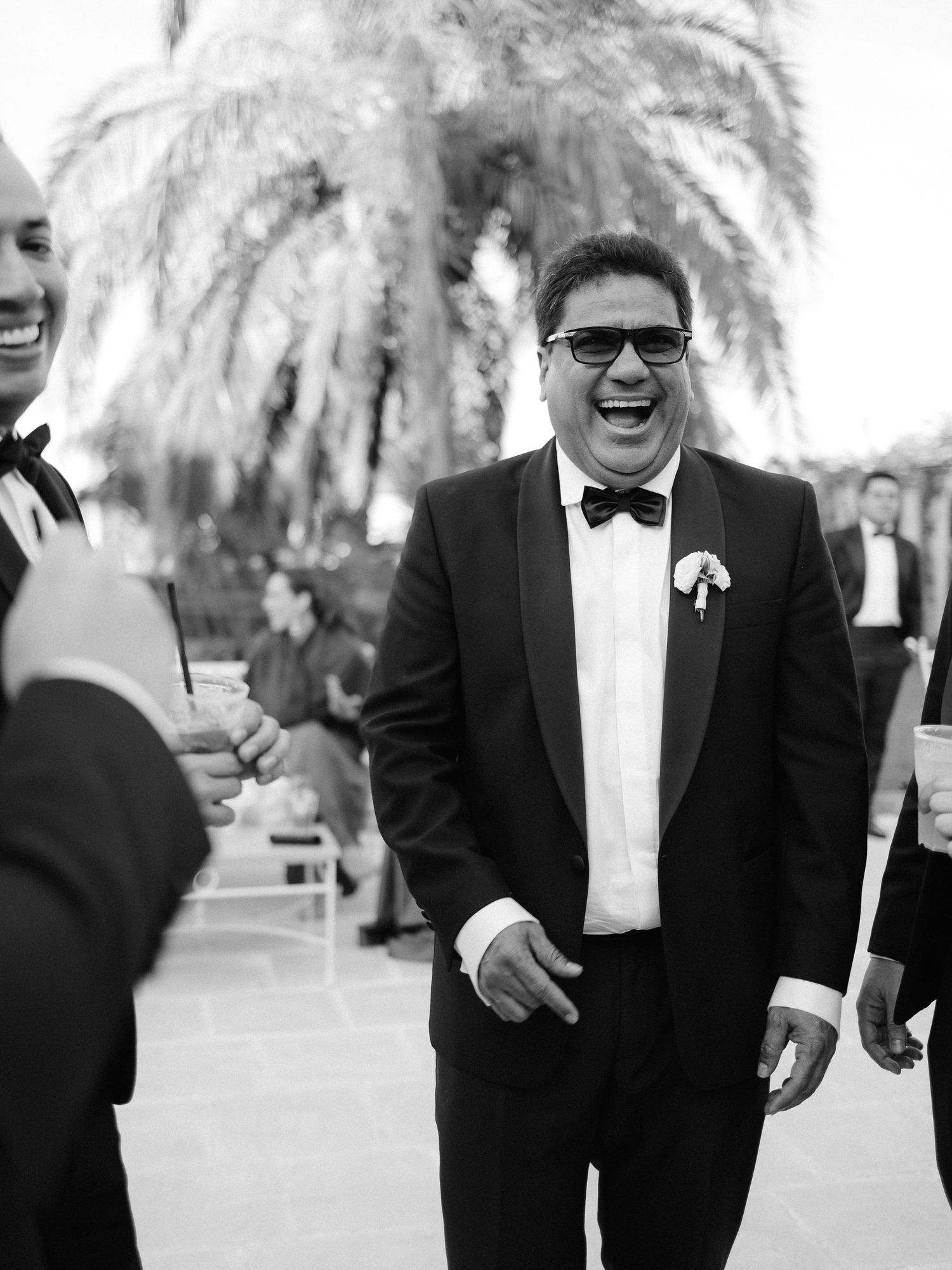 Black and white photo of a joyful person in a tuxedo laughing heartily at an outdoor wedding celebration with a palm tree behind.