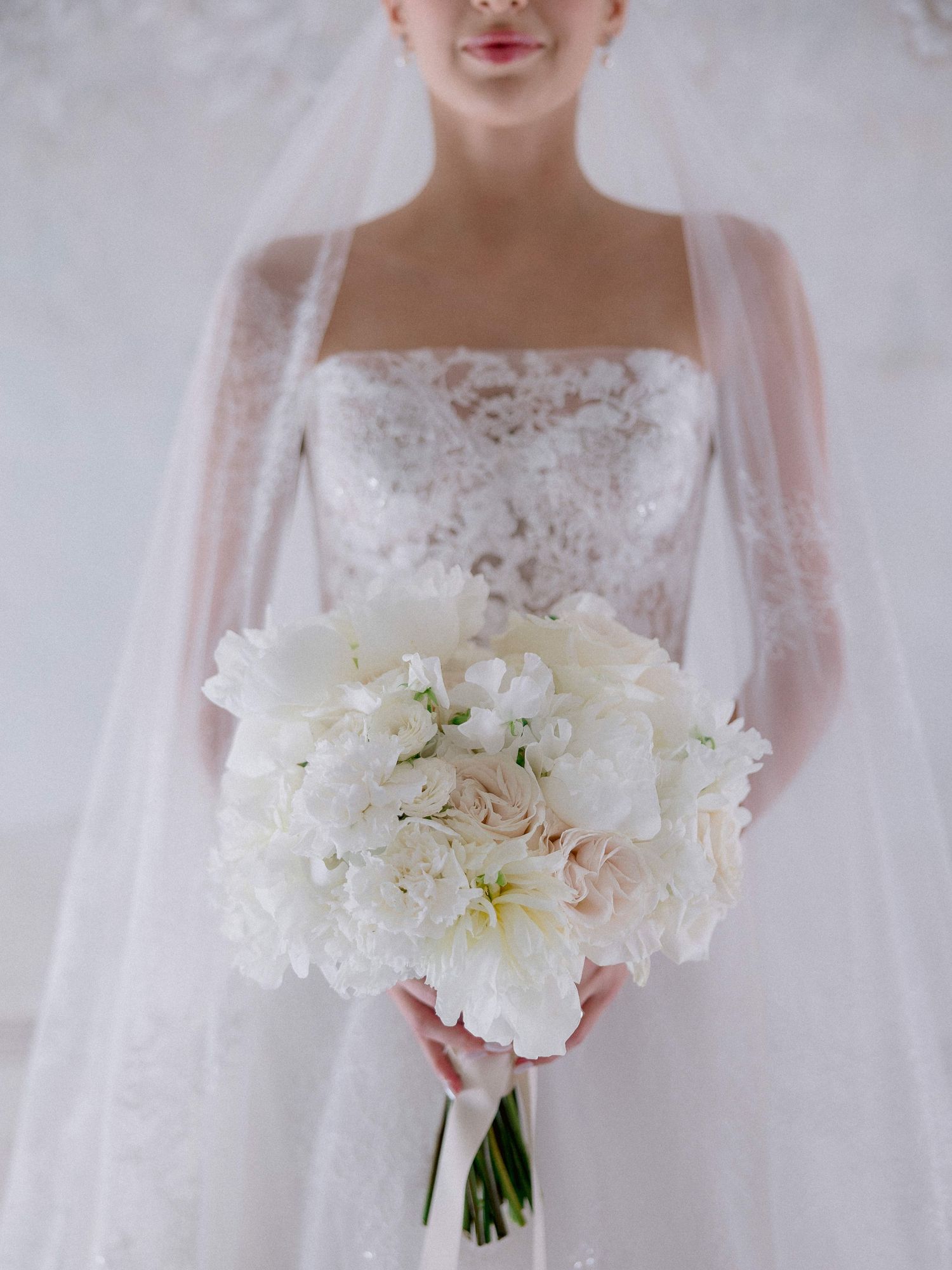 Elegant white bridal bouquet with lilies and peonies held against a lace wedding dress with sheer tulle veil.