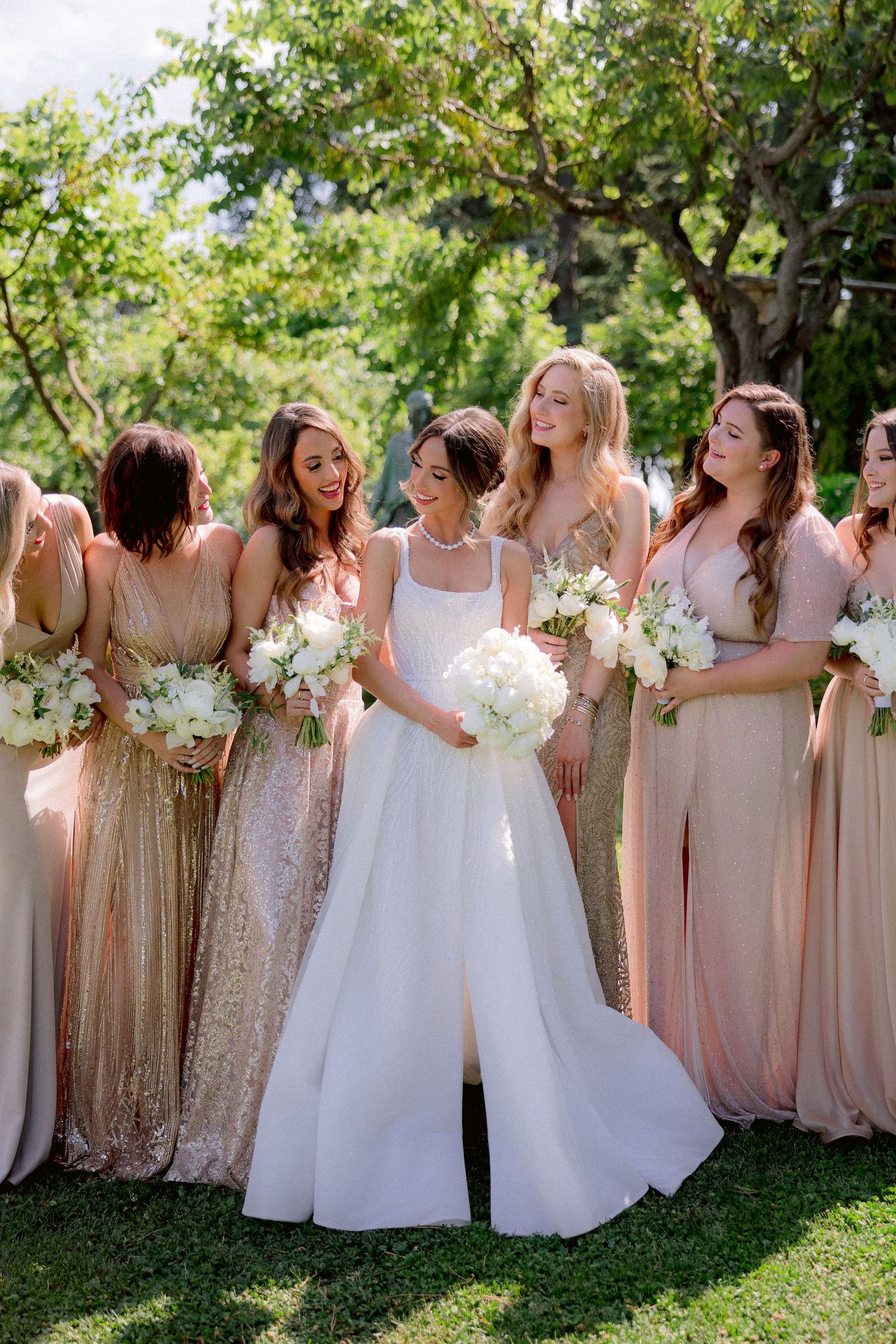 Bridesmaids in blush and gold sequin dresses laugh together with bride in white gown while holding floral bouquets outdoors.