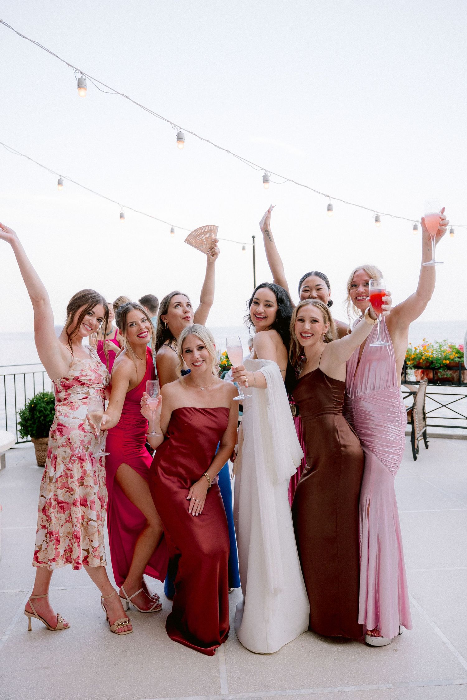 A celebratory group of women in elegant dresses pose together at an outdoor wedding reception with string lights above.