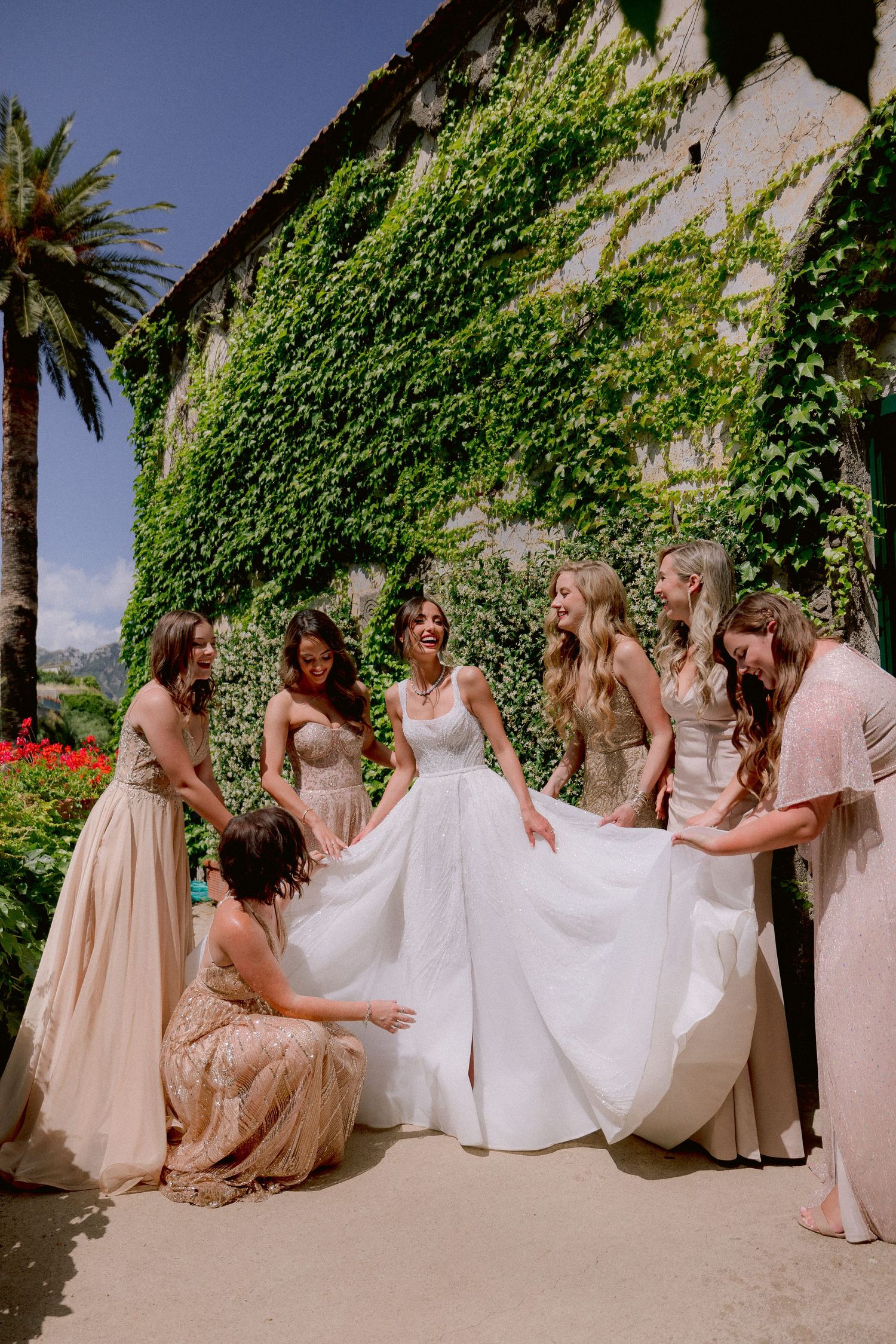 Bridesmaids in blush pink dresses help the bride with her flowing white wedding gown against an ivy-covered wall.