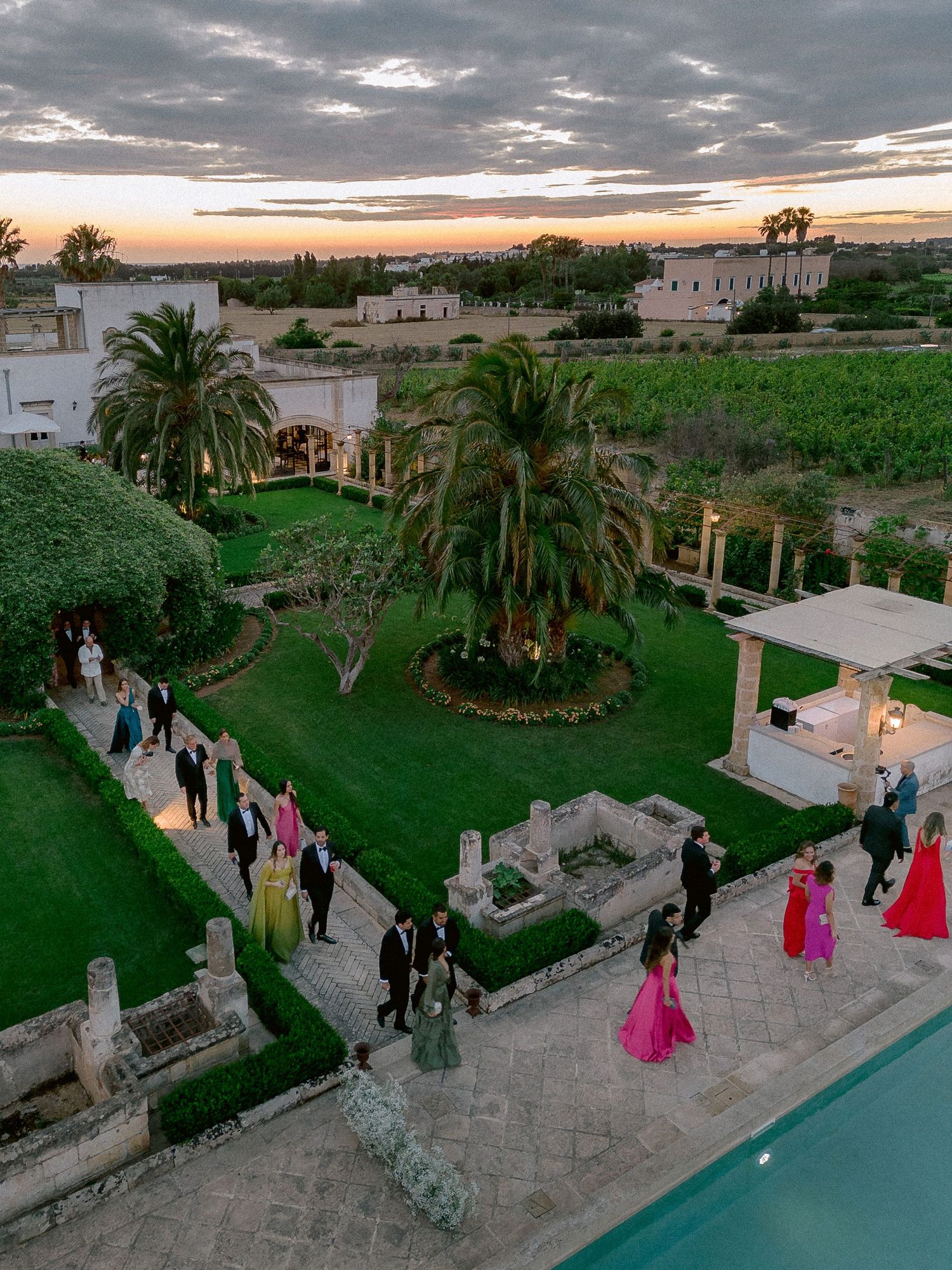 Elegant outdoor event at sunset with guests gathered on a stone patio overlooking a pool and lush garden with palm trees in Puglia.