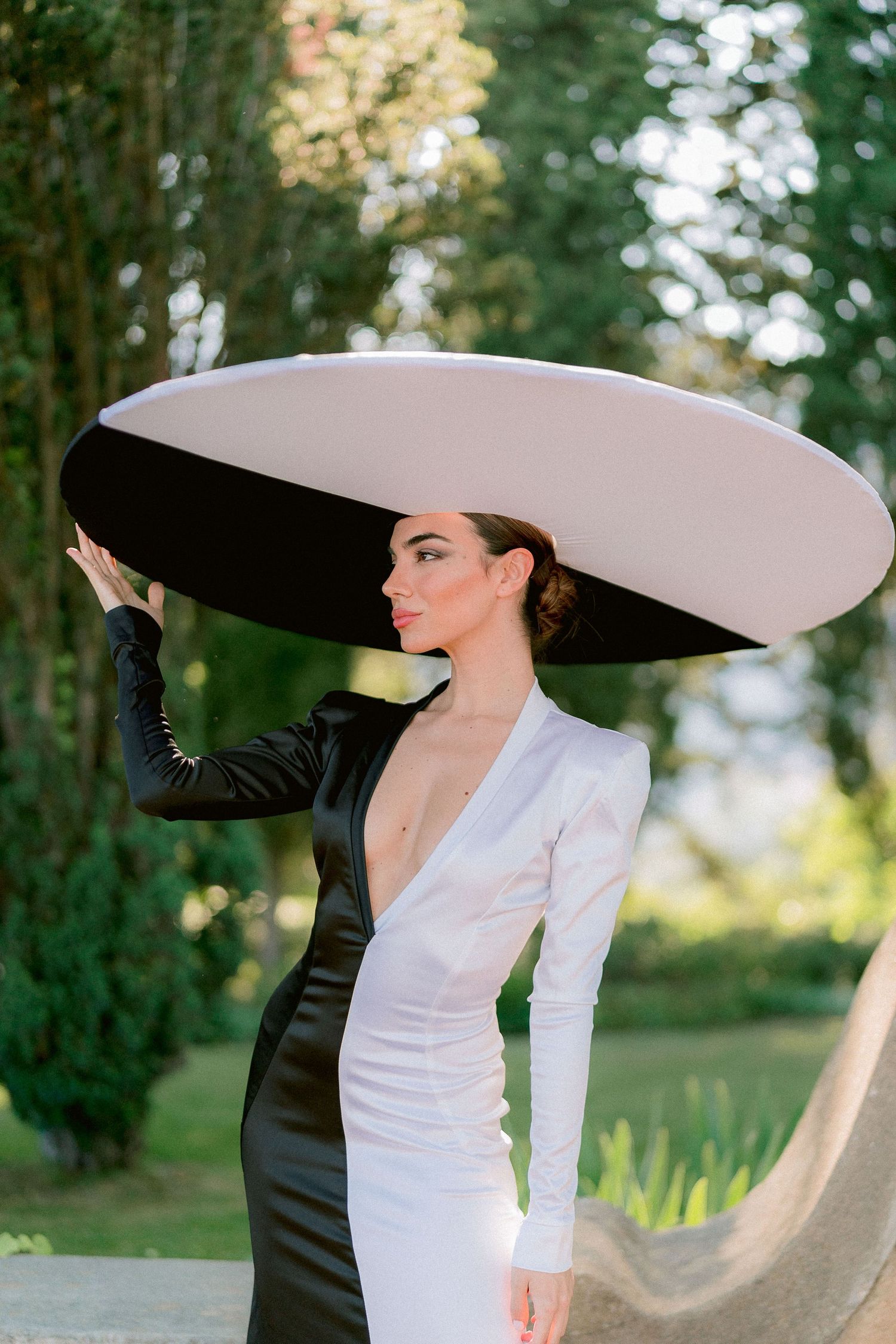 A model in a dramatic black and white colorblock dress poses outdoors wearing an oversized wide-brimmed white hat.