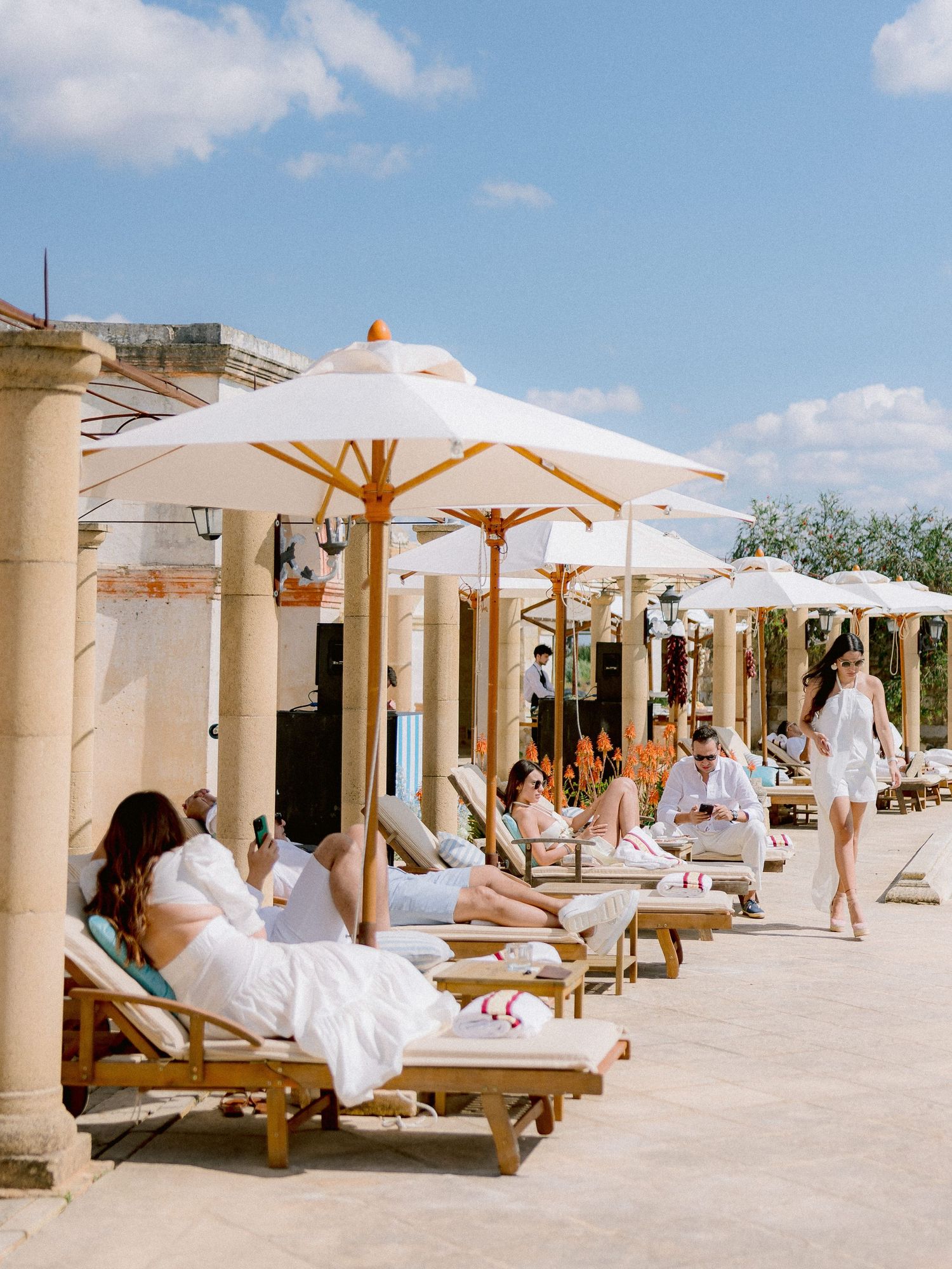 Guests relax on wooden lounge chairs under white umbrellas at a luxury resort patio on a sunny day.