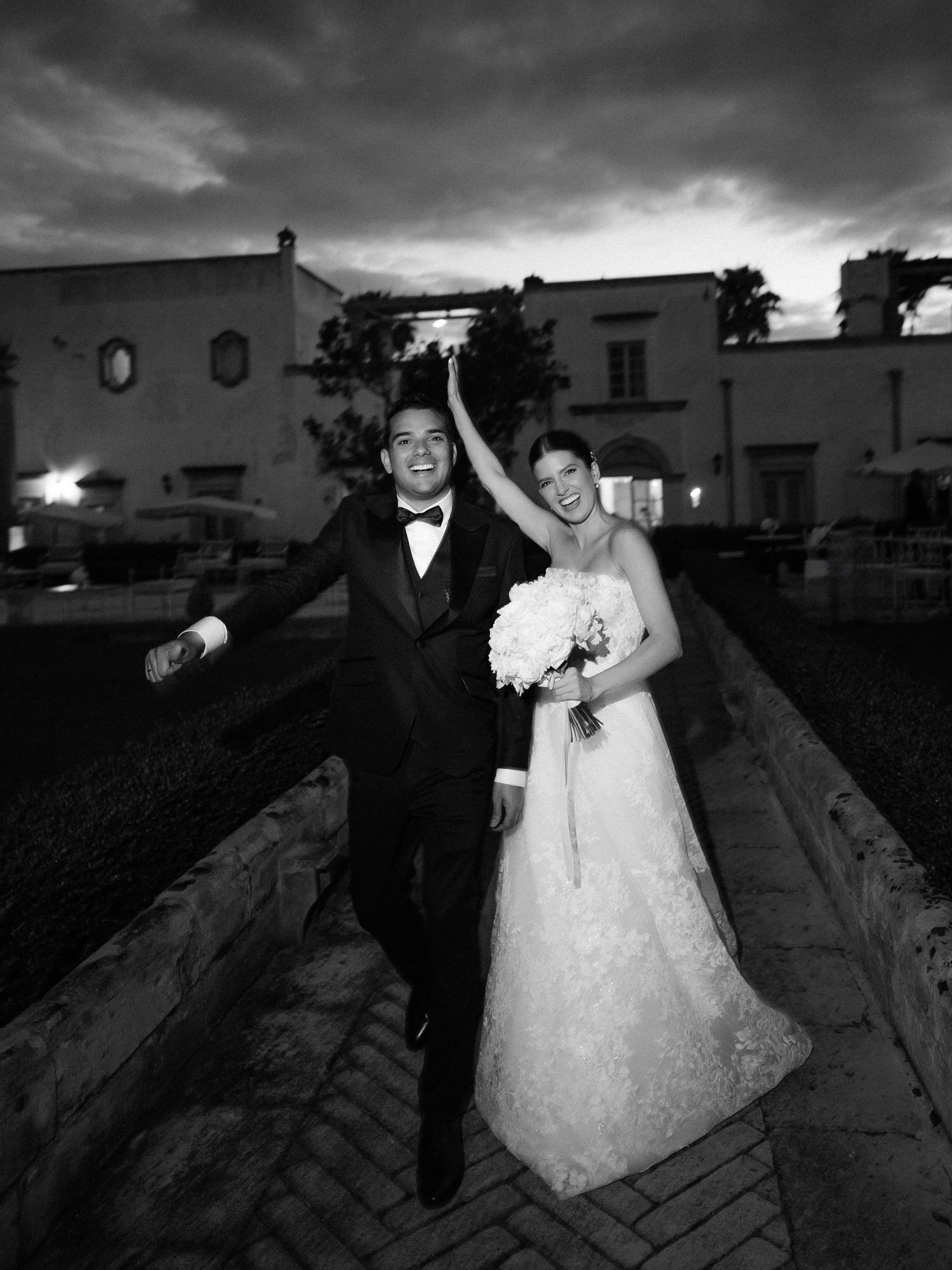 A wedding couple poses joyfully on a brick path at dusk with dramatic storm clouds in the background.