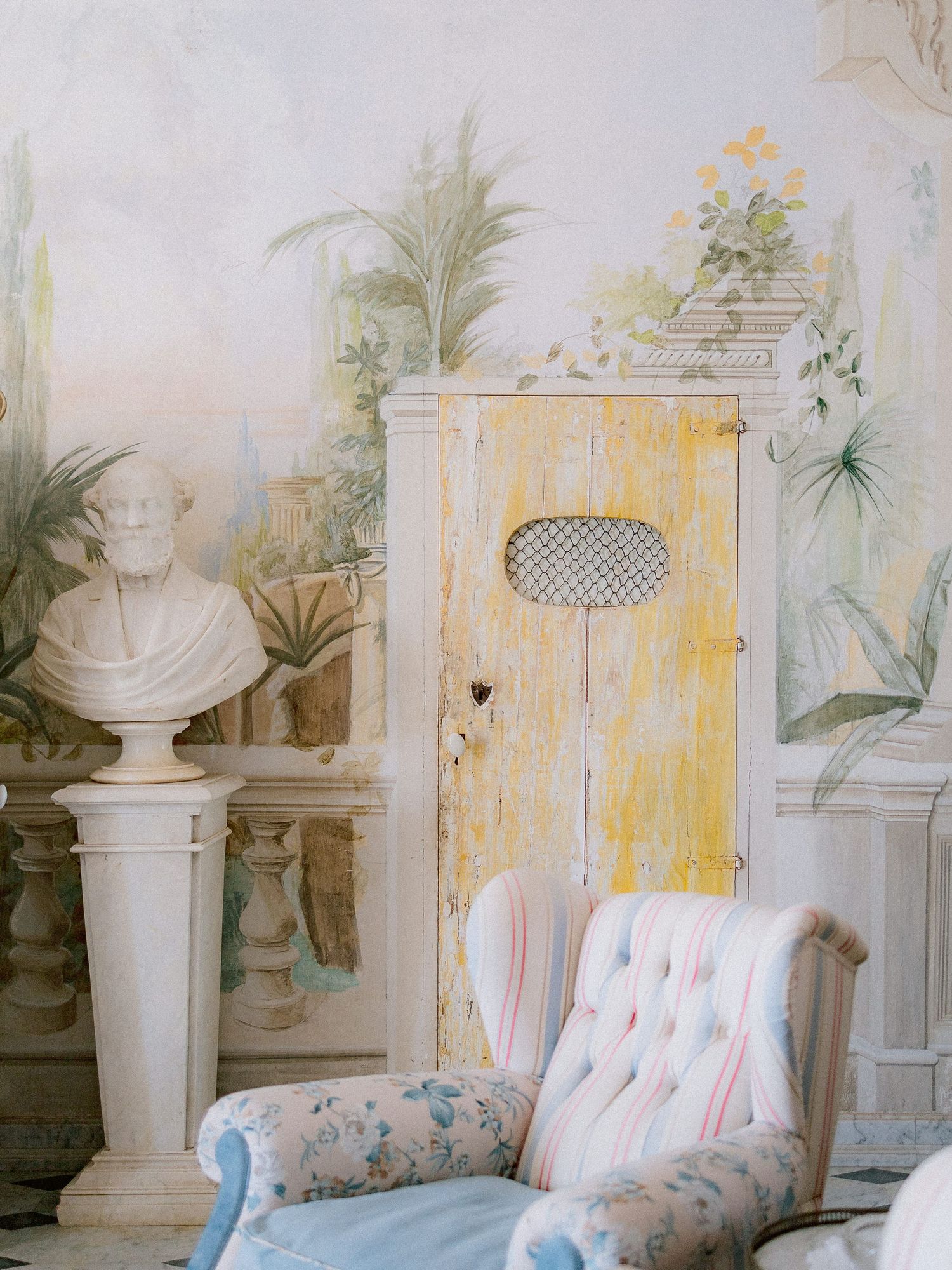 Vintage floral armchair and yellow wooden cabinet against pastel wall mural with classical bust sculpture decoration.