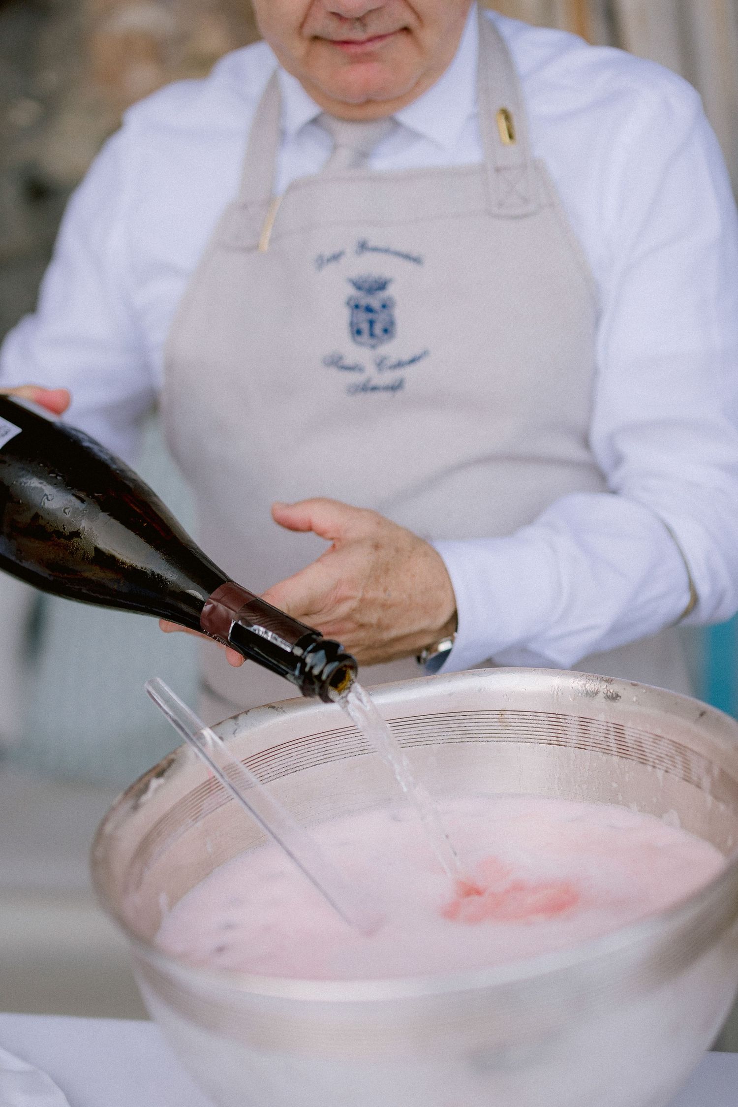 Someone wearing a gray apron pours wine into a bowl of pink-colored mixture from a dark bottle.