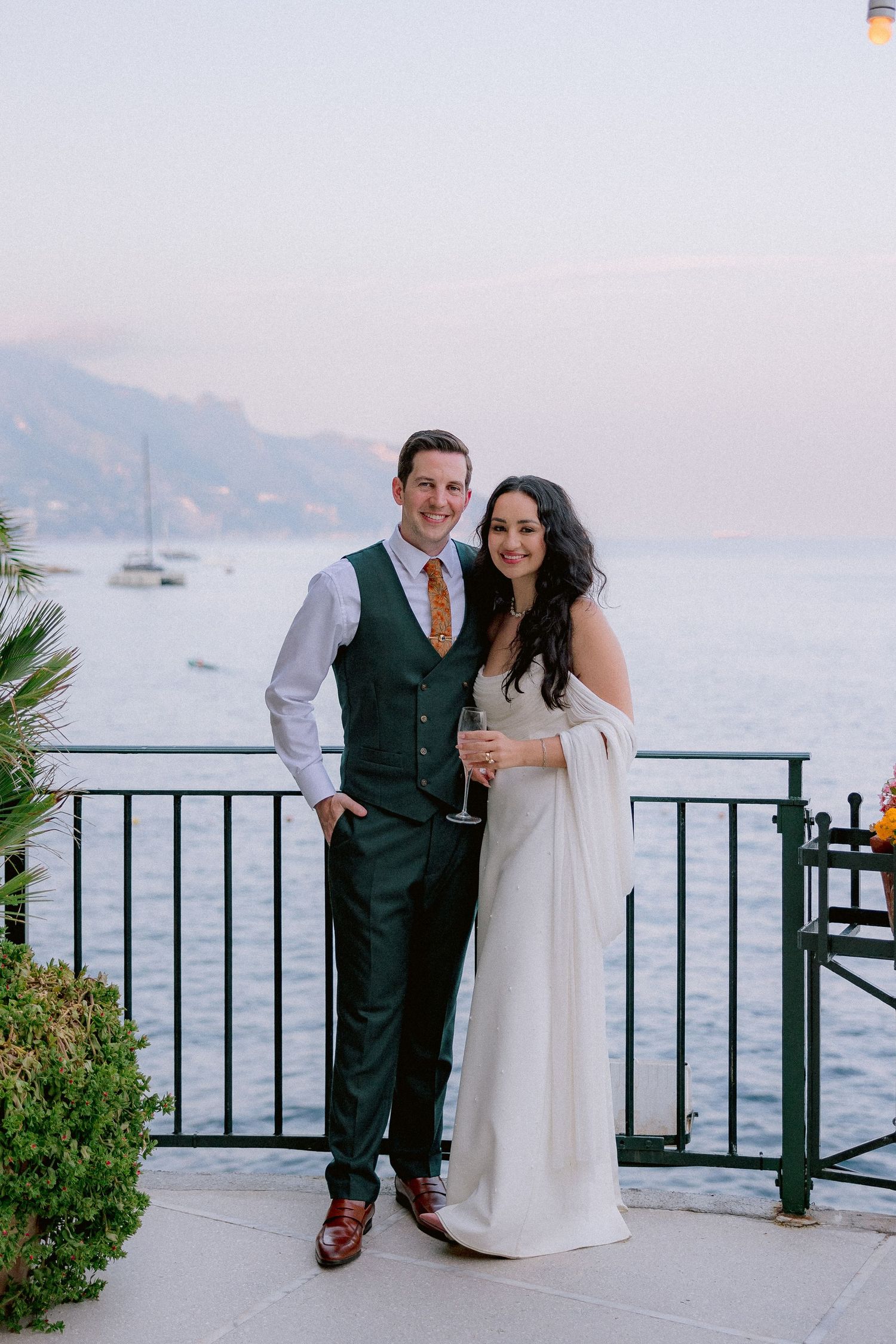 A couple in formal attire poses on an oceanfront terrace with boats and mountains visible in the background.