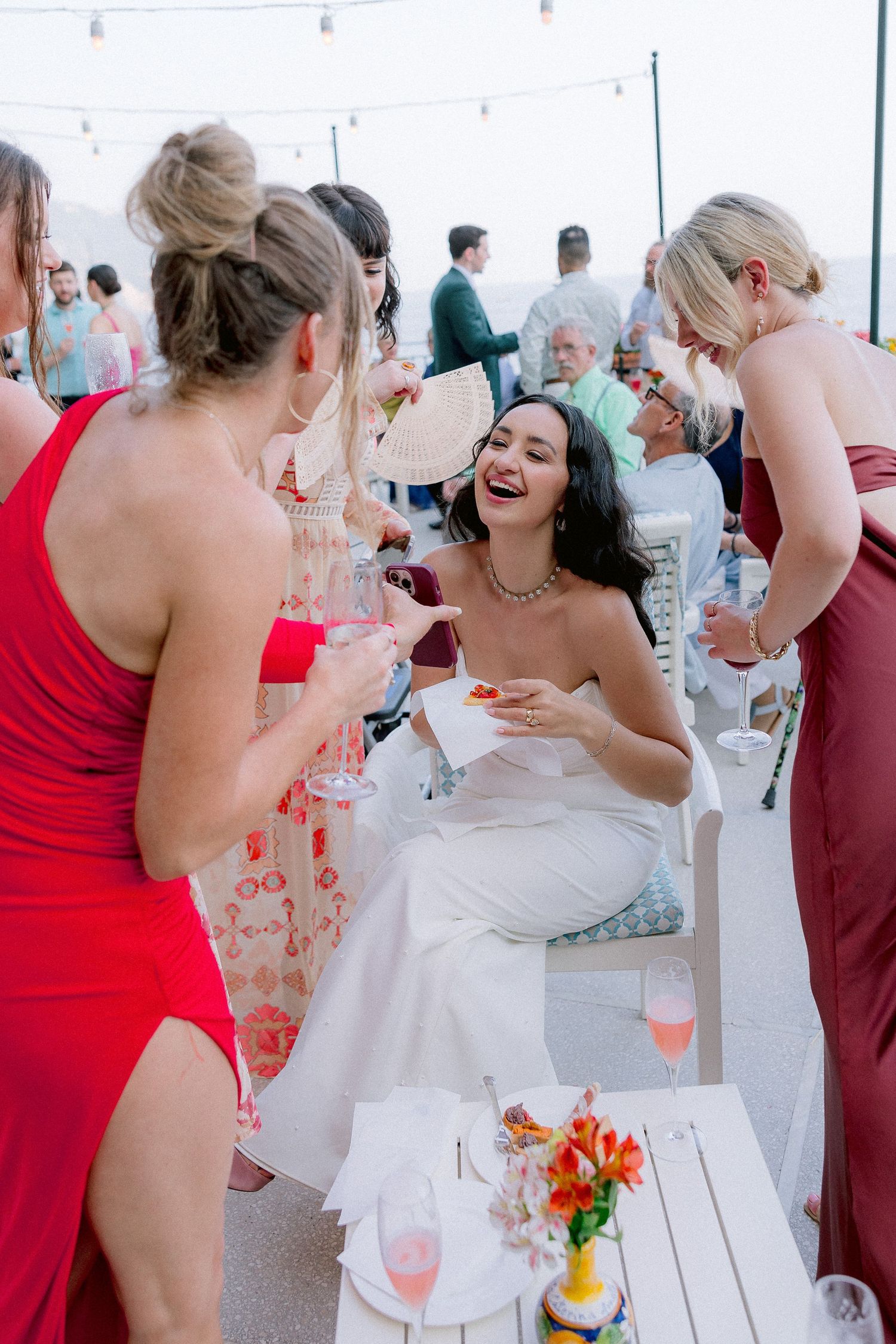 A celebration at an outdoor reception with people in red dresses and a white wedding gown laughing together.