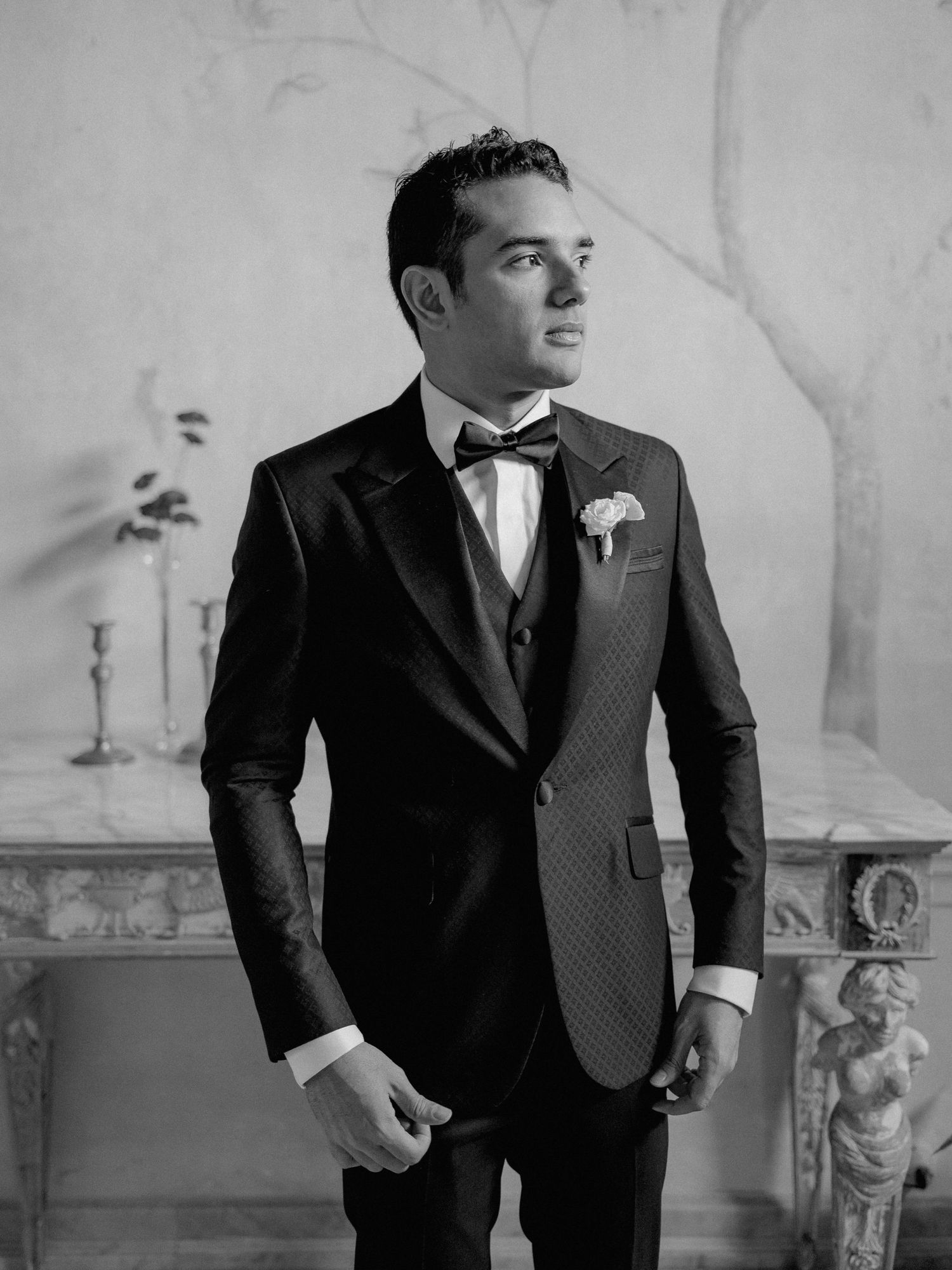 A groom in a classic black tuxedo with bowtie and boutonniere looking to the side in an elegant indoor setting.