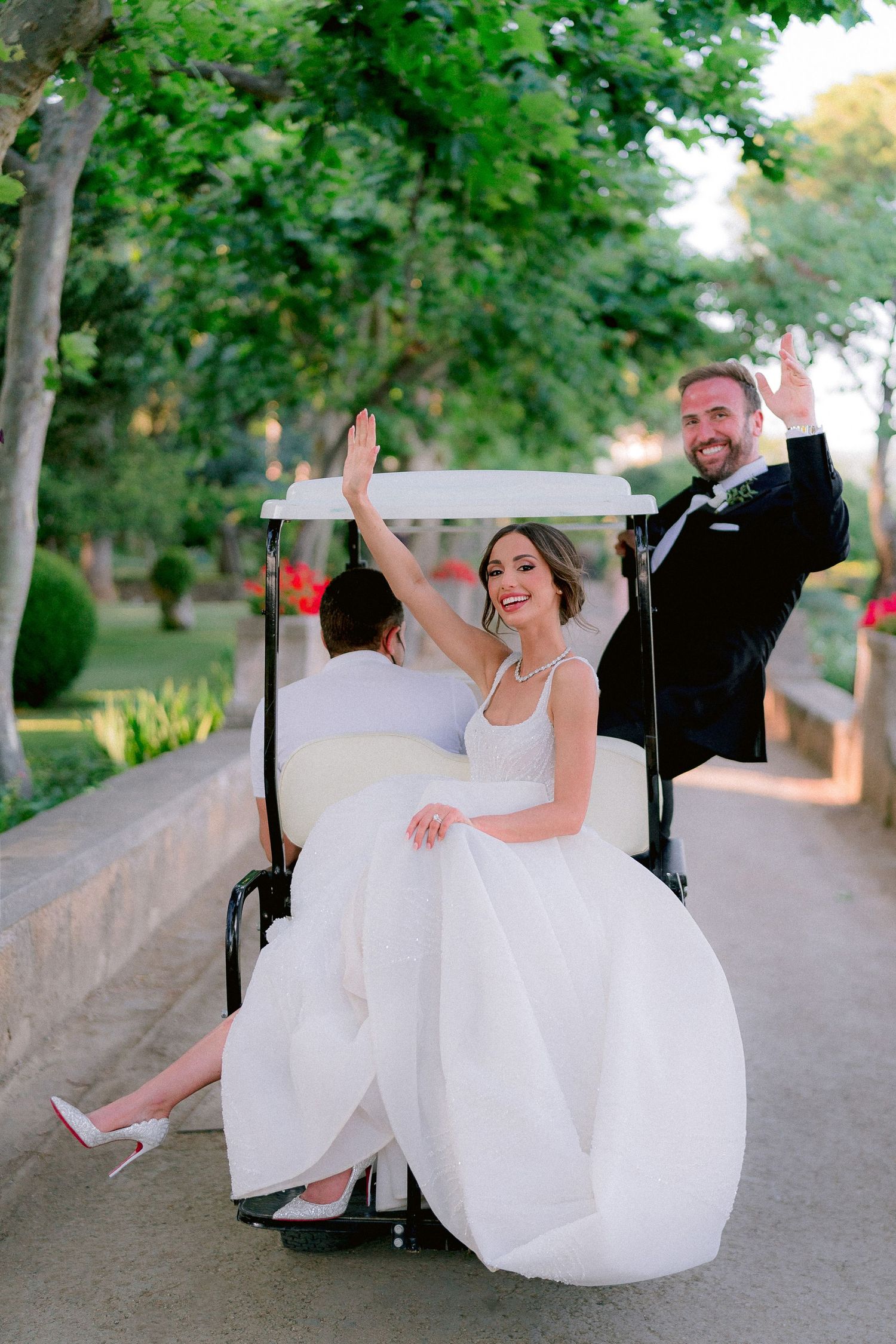 Newlywed couple riding in a golf cart along tree-lined path as bride in white gown waves her hand joyfully.
