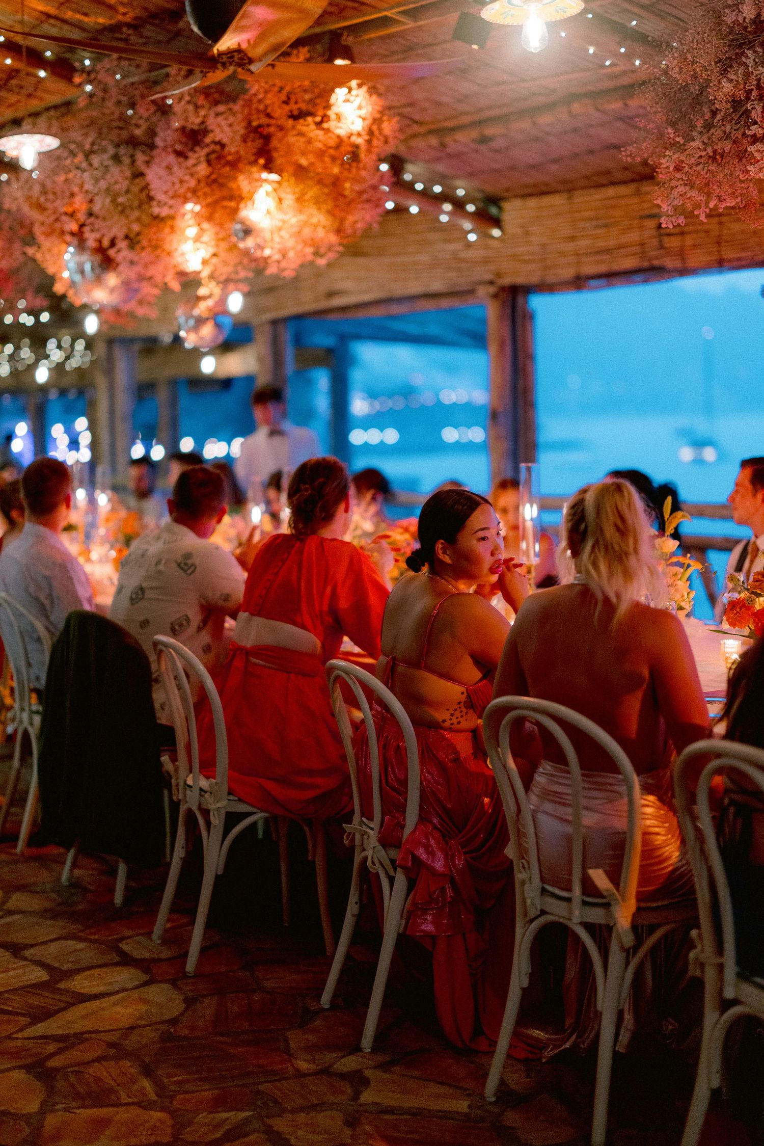 Diners enjoy an evening meal at a waterfront restaurant with string lights and pink floral decorations overhead.